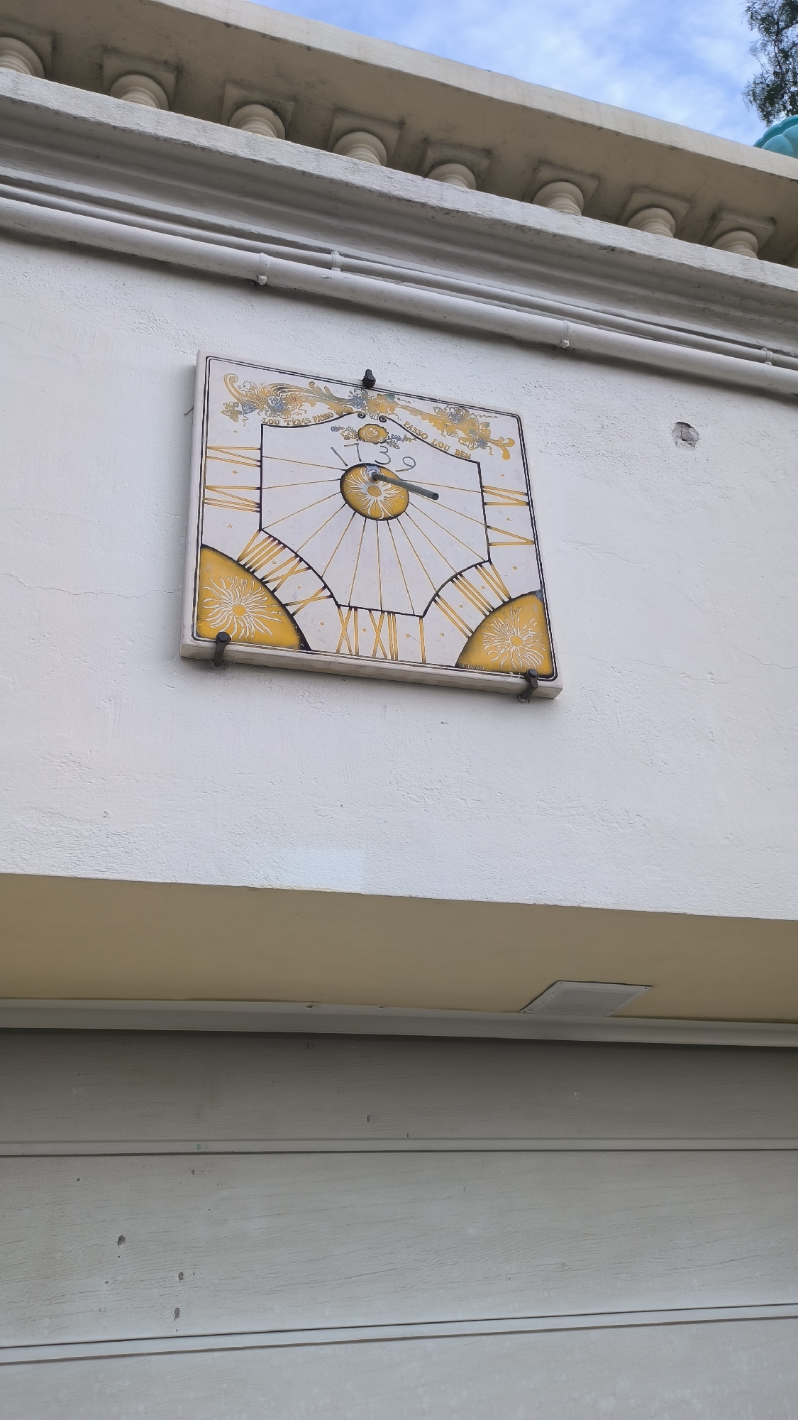 A square sundial, attached to a white stucco wall, is the primary subject. The sundial features Roman numerals and decorative yellow and blue patterns. A metal gnomon, the part of the sundial that casts the shadow, is visible. Above the sundial, a section of a building with columns and decorative trim is present. The sky, with some clouds, is visible in the upper portion of the image. Below the sundial, a beige overhang and a grey horizontal siding material are visible. The overall scene appears to be outdoors.