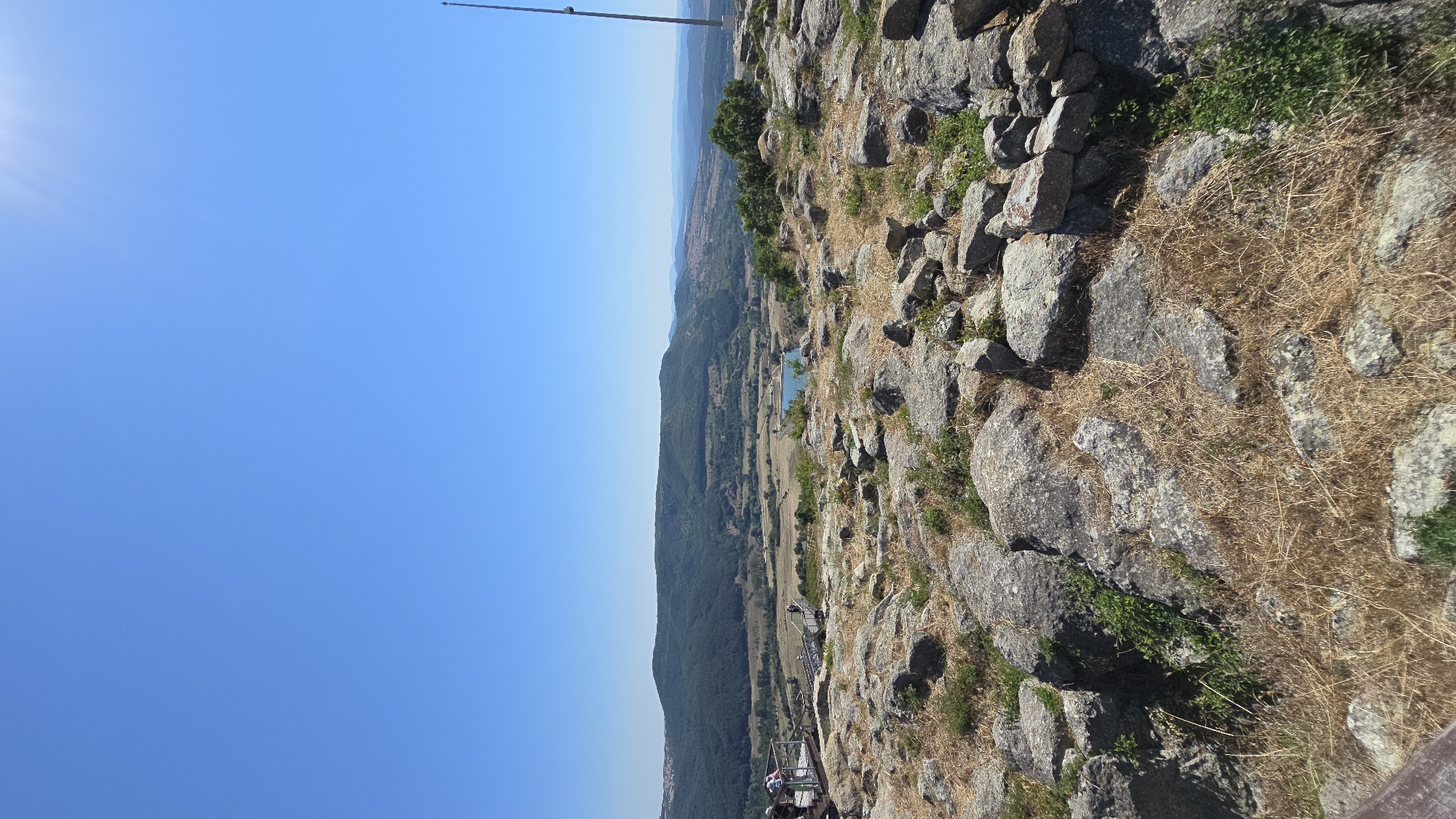 The image depicts a panoramic view from a rocky, elevated terrain overlooking a valley and distant hills. The foreground is dominated by large, grey rocks and dry, yellowish grass, suggesting a somewhat arid environment. In the middle ground, there are wooden walkways and viewing platforms with several people observing the landscape. Beyond the platforms, the terrain slopes down into a valley with patches of greenery, fields, and a small, blue body of water. Further in the distance, rolling green hills with scattered trees form the backdrop, under a clear, bright blue sky. The sun is visible in the upper right corner, casting a bright glare. A tall, thin antenna or tower stands on the right edge of the frame, also on the rocky hilltop. The scene appears to be a historical or natural site, possibly a fortification or ancient ruins, given the presence of the viewing platforms and the rough terrain. The location is identified as Gorna Krepost, Bulgaria.