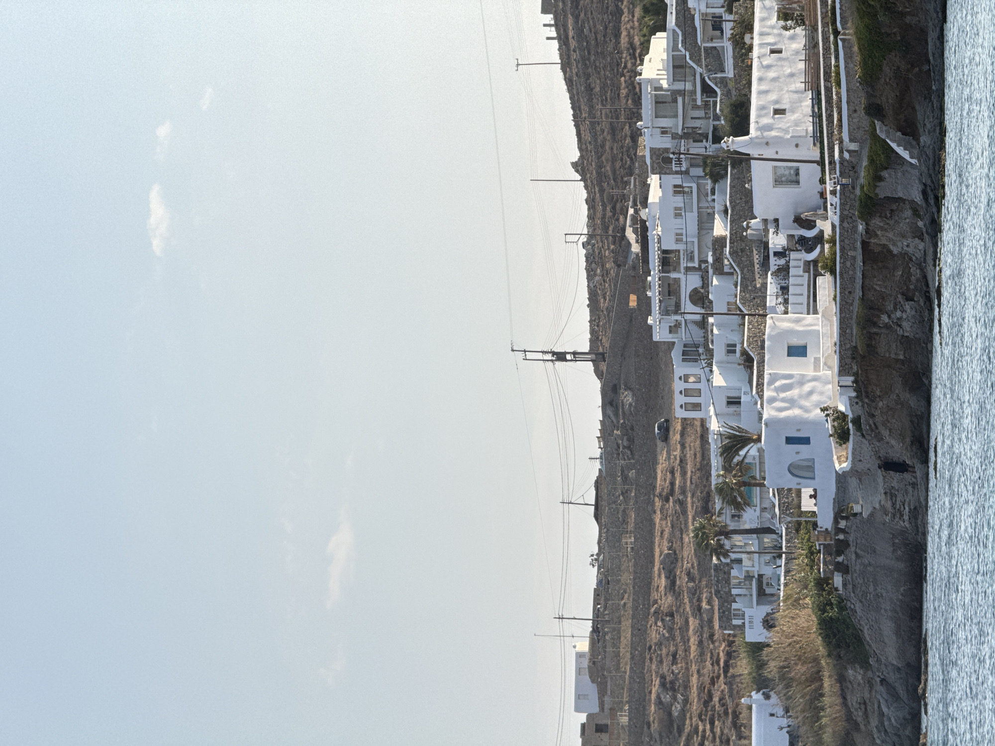 The image shows a view of the coastal town of Agios Ioannis Diakoftis, Greece, with white buildings scattered on a hillside overlooking the Aegean Sea. The architecture is characteristic of the Greek islands, with whitewashed cubic houses featuring blue accents. In the foreground, the calm blue water of the sea laps against a rocky shore. A single person in dark clothing is visible standing on a path leading up from the water. The hillside is dotted with sparse vegetation, rocks, and some palm trees. Power lines stretch across the landscape, supported by utility poles. The sky is a pale blue with a few wispy clouds. The overall scene is serene and picturesque, typical of a Mediterranean coastal village.