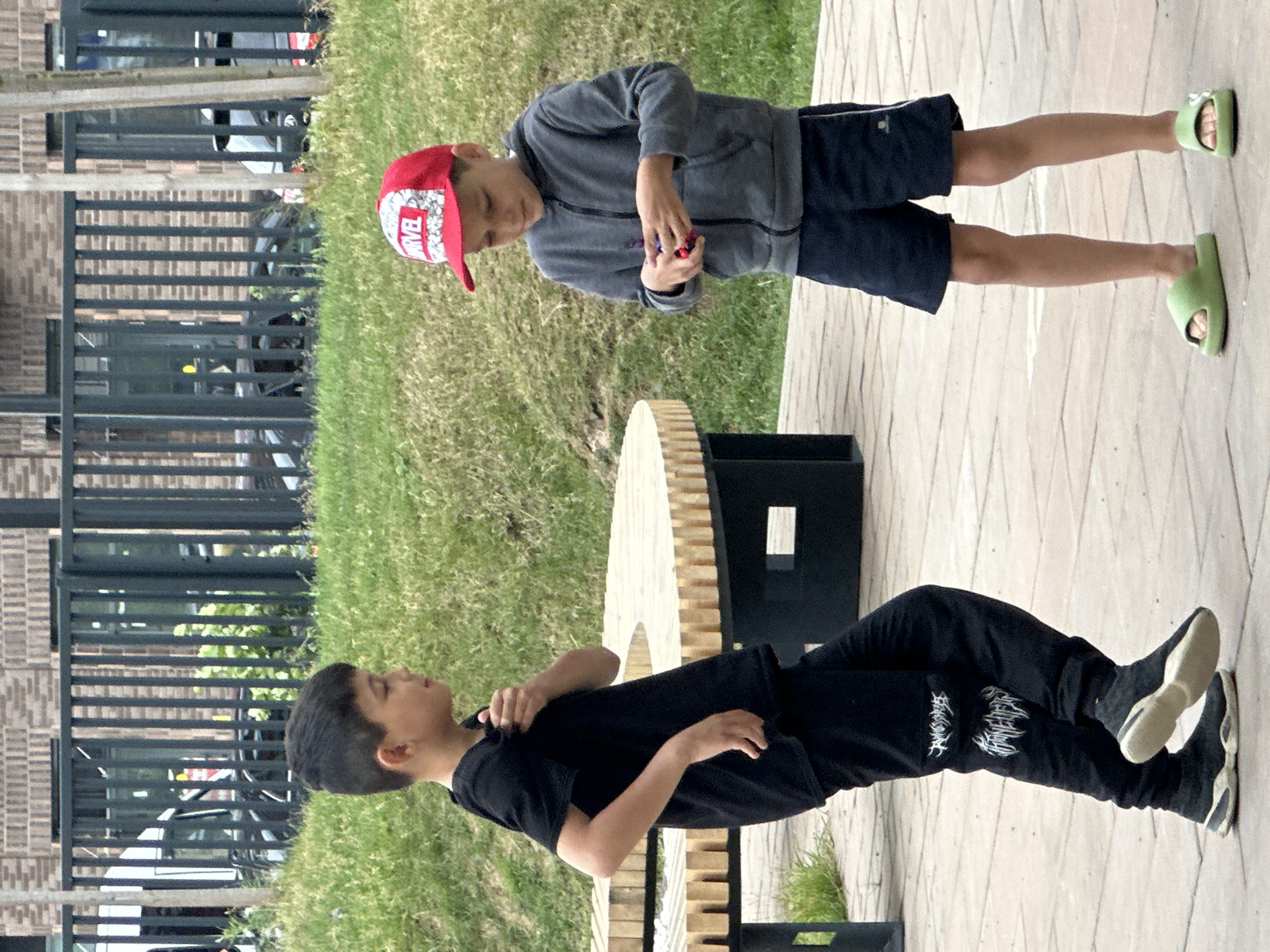 Two young boys are standing in an urban outdoor area with a paved ground surface and grassy sections. The boy on the left, wearing a black t-shirt and black pants with white graphic designs, is looking to his right with his right hand touching his shirt. The boy on the right, wearing a grey hooded jacket, navy shorts, and a red Marvel-branded baseball cap, is looking down at his hands, which are holding a small red object. In the background, there is a circular wooden table with dark metal legs and a metal fence in front of a brick building. The scene is likely located in Znamya Oktyabrya, Russia.
