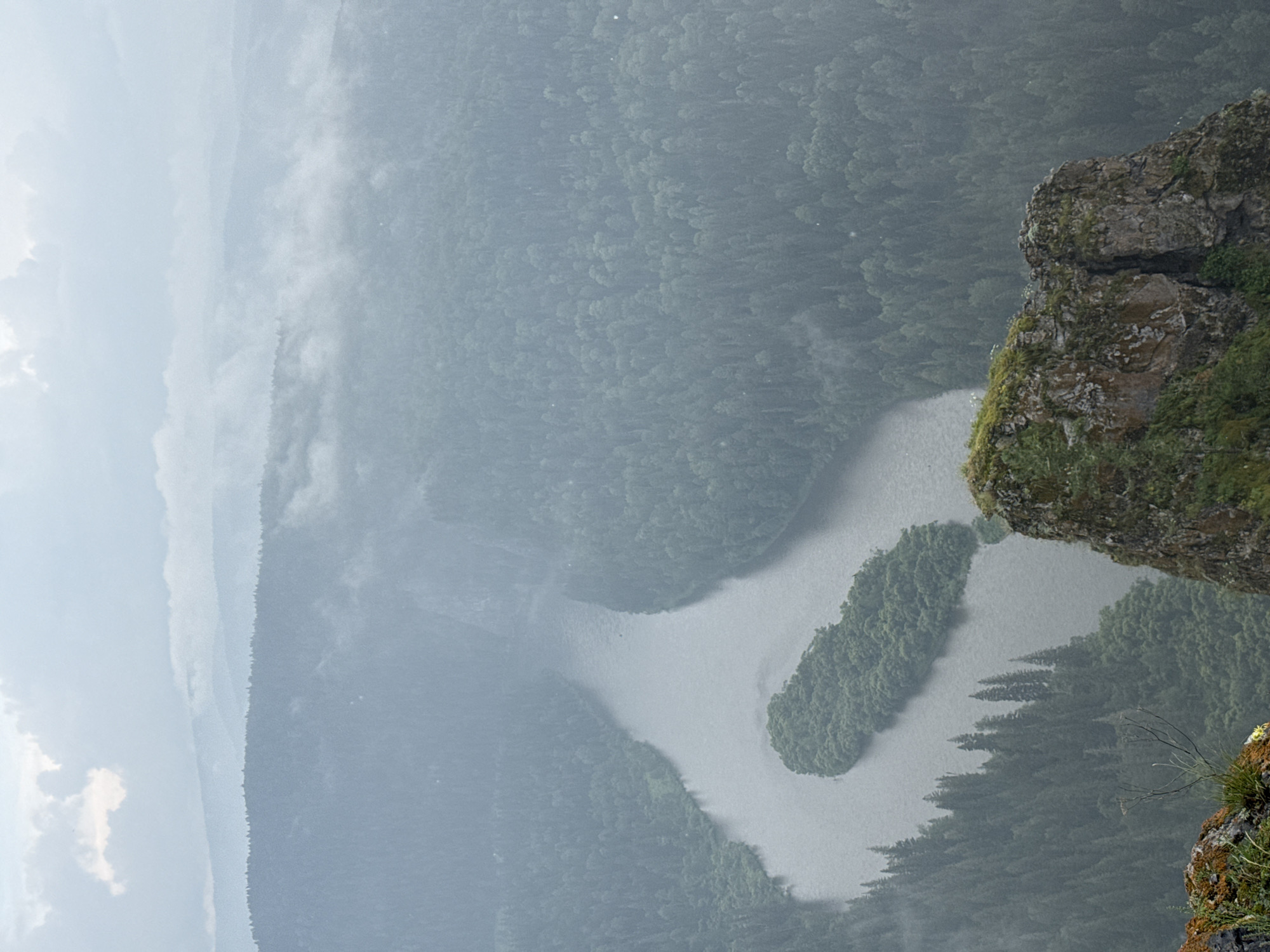 The image captures a panoramic view of a winding river flowing through a dense forest in the city of Urman, Russia. The landscape is characterized by rolling hills covered in evergreen trees, with a prominent island situated in the middle of the river. The sky is overcast with clouds, and a light mist hangs over the distant hills, creating a hazy, atmospheric effect. In the foreground, a rocky outcrop with sparse vegetation overlooks the river valley. The scene conveys a sense of natural beauty and tranquility.