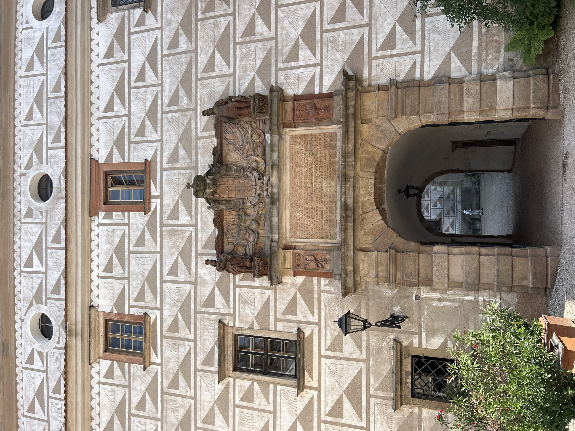 This is a medium-angle, eye-level shot of the Sgraffito courtyard entrance at Náchod Castle in Náchod, Czechia. The facade is adorned with a repeating geometric sgraffito pattern in shades of white and beige. An arched stone entryway dominates the lower center of the frame, leading to a courtyard visible in the background. Above the arch, a large, ornate stone crest with a crown and figures is mounted on the wall. Two stone statues stand on either side of the crest. A large inscription is carved into a stone panel below the crest. To the left of the arch, a tall, dark lantern with a decorative bracket is attached to the wall, positioned above a window with a black iron grate. A lush green plant in a planter sits in the lower left corner of the image. The overall impression is one of historical architecture and intricate design.