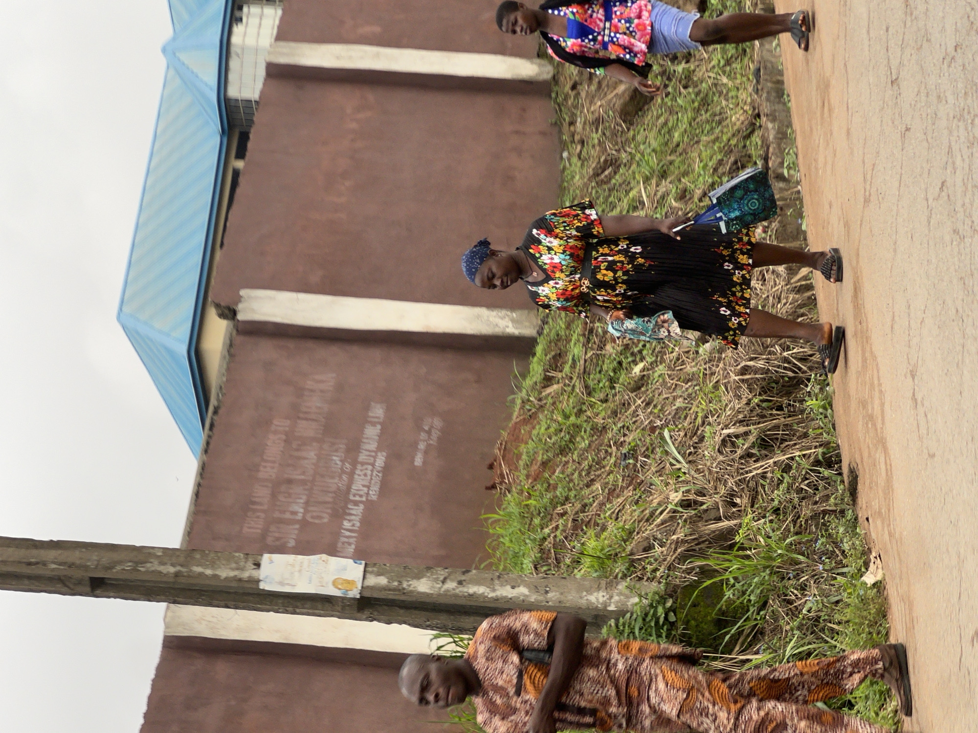 In Nkpor, Nigeria, a street scene unfolds with a man and two women walking along a dusty road. The man on the left, clad in a brown and orange patterned dashiki, pauses near a concrete utility pole. Behind him, a wall with faded inscriptions, including "THIS LAND BELONGS TO SIR ENGR. ISAAC NWABUEKE ONWUEGBUSI" and "MEXY ISAAC EXPRESS DYNAMIC LINK RENDEZVOUS," suggests a property boundary.
To the right of the utility pole, a woman in a black dress adorned with colorful floral patterns walks, her head covered with a blue bandana. She carries a patterned shopping bag and a folded cloth. Further to the right, a younger woman in a vibrant floral-patterned top and a light blue skirt walks ahead. The environment is characterized by overgrown grass and a cloudy sky, with a light blue-roofed building visible in the background. The overall impression is one of everyday life on a Nigerian street.