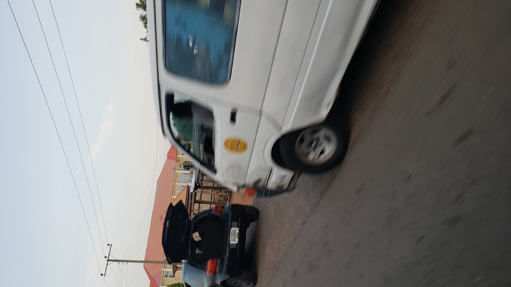 In Jalingo, Nigeria, a white van is captured in motion on a paved road. The van's front is in the foreground and slightly blurred due to movement. A yellow circular emblem is visible on the van's side. To the left of the van, a black hatchback car is parked with its trunk open. In the background, buildings with orange-tiled roofs are visible, along with a utility pole with electrical wires. The sky is overcast, and the overall lighting suggests it is either early morning or late afternoon.
