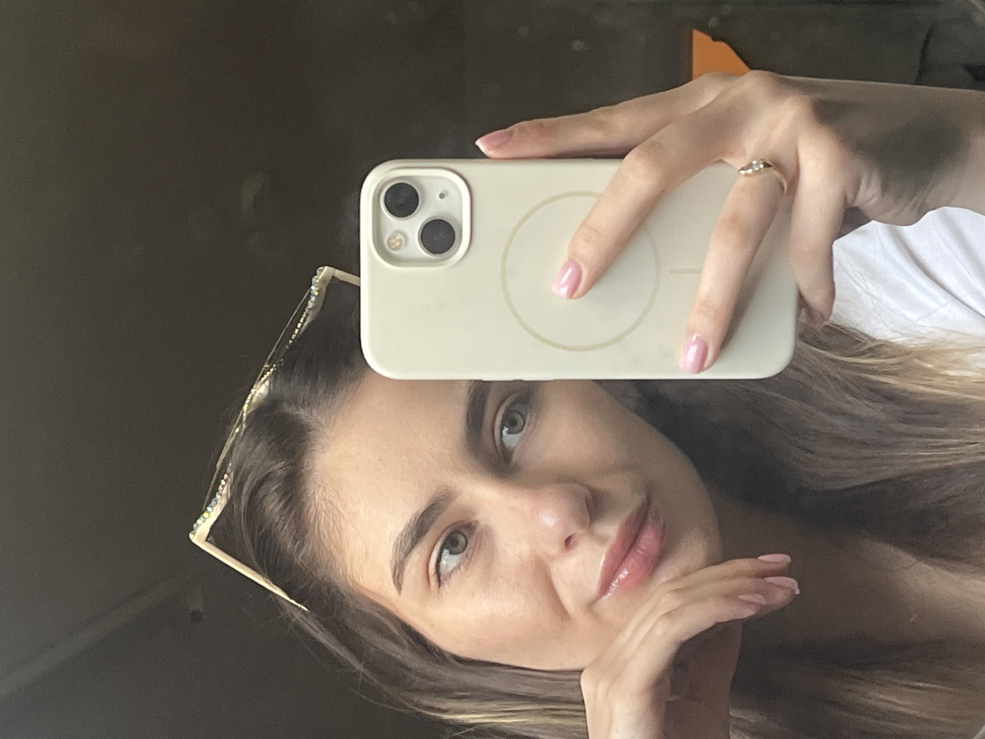 A woman is taking a selfie in a mirror. She is holding a white iPhone 13, and a pair of sunglasses with rhinestones are perched on top of her head. She is wearing a ring on her ring finger, and her nails are painted pink. The background is blurry, but appears to be a room with a dark-colored wall. Location: Unknown, Unknown.