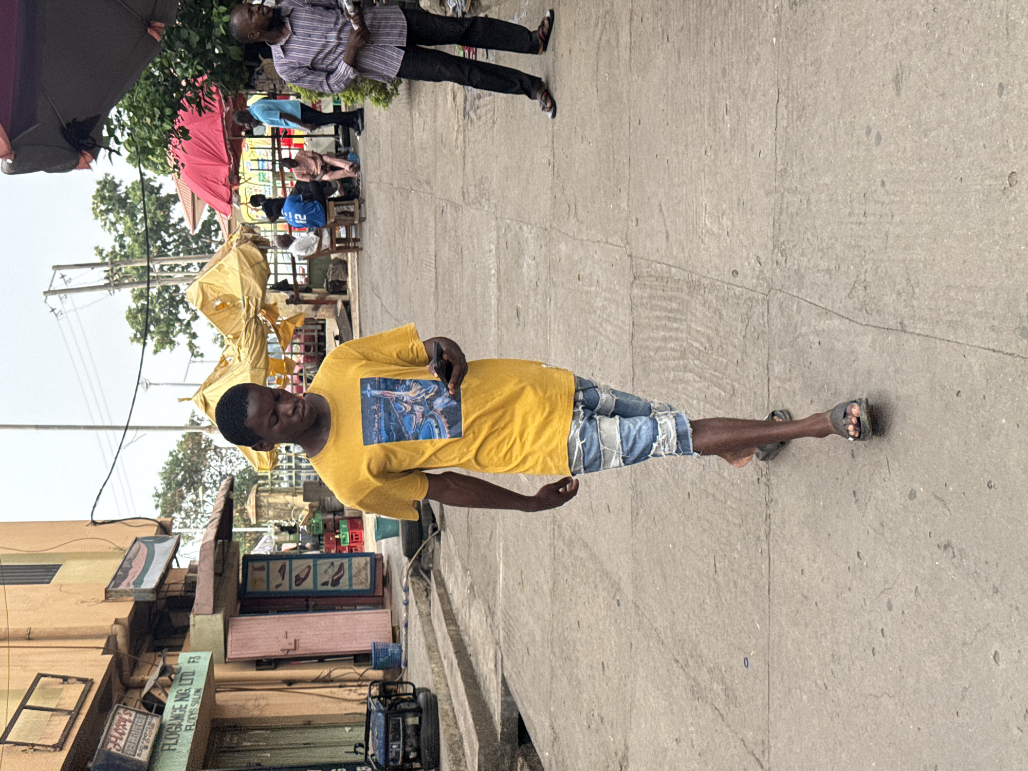 A man is walking along a sidewalk in Abuja, Nigeria. The man is wearing a yellow t-shirt and cutoff denim shorts and is wearing sandals. Another man walks behind him. A series of street vendors with umbrellas are in the background. Some buildings are on the other side of the sidewalk.