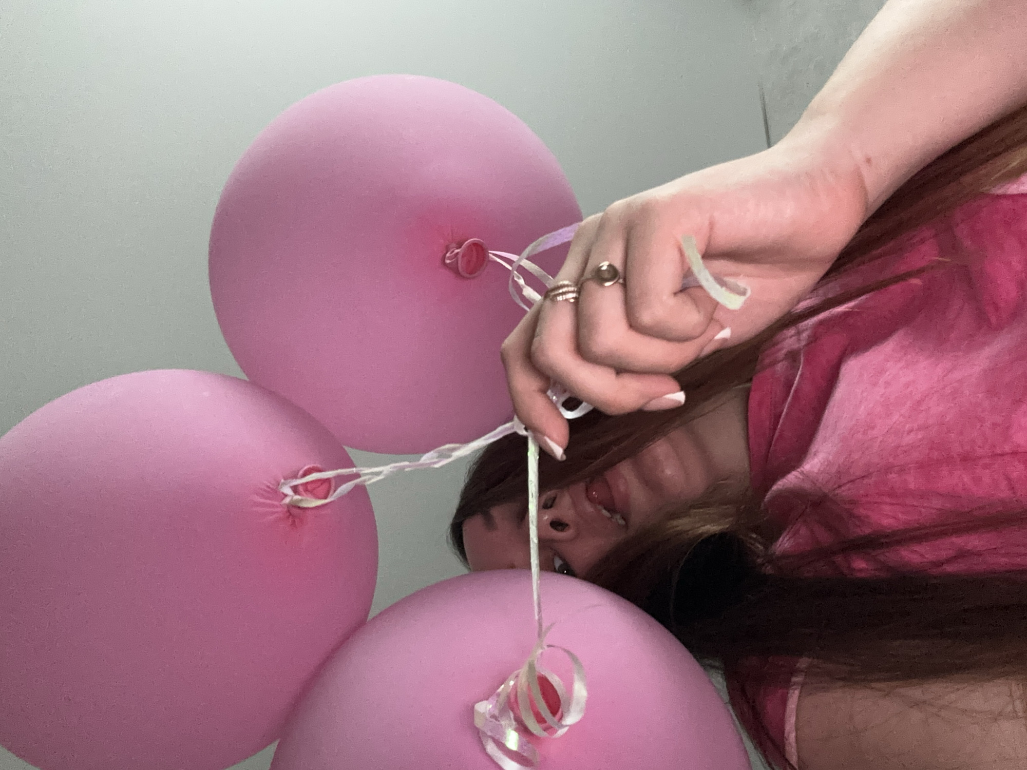 The image shows a woman holding three pink balloons. She wears a pink shirt and several rings on her fingers. The background is a light gray wall. The location, Chaikovsky, Russia, cannot be confirmed from the image alone.