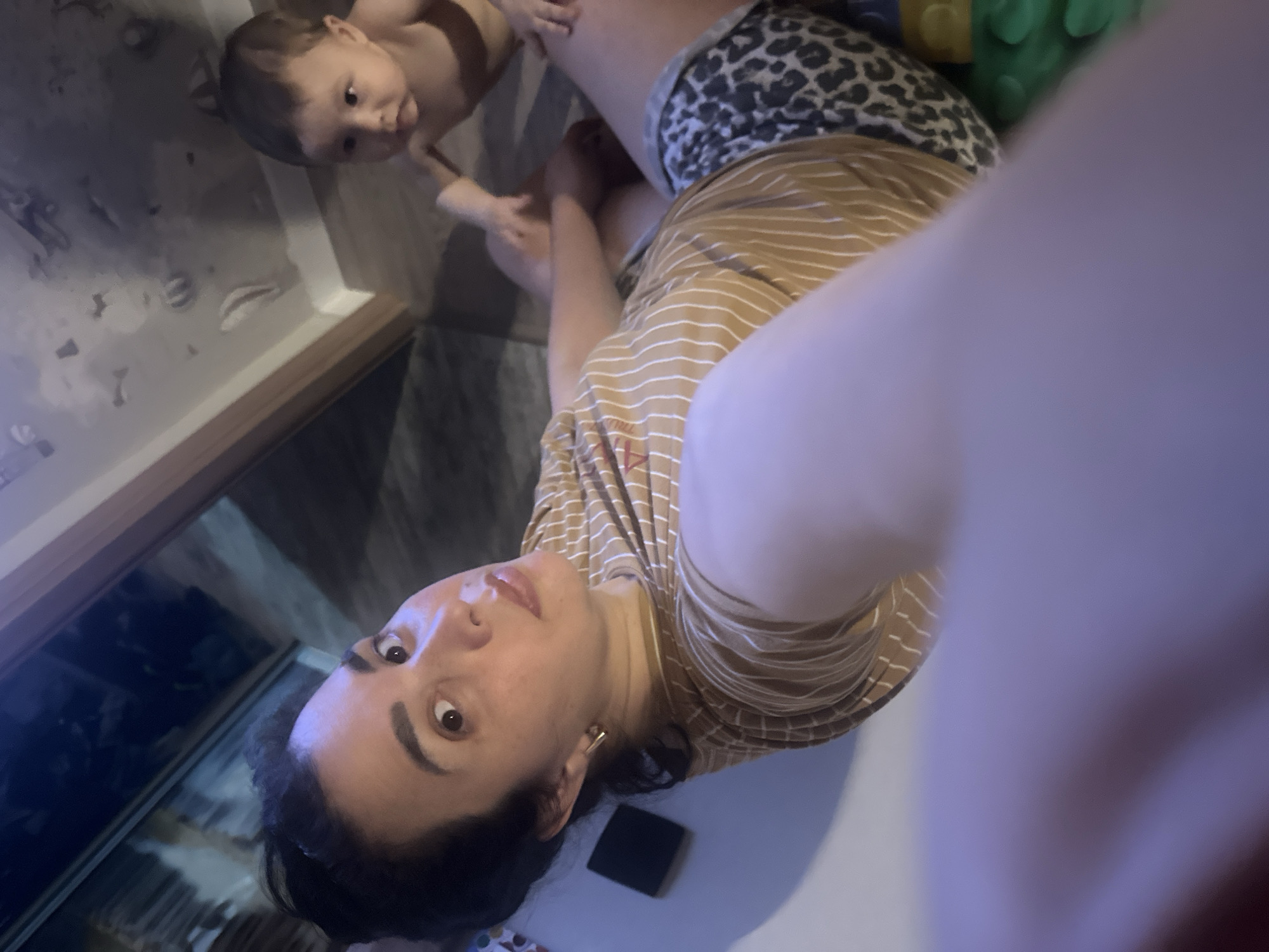 An overhead shot shows a woman in a tan and white striped t-shirt and leopard-print shorts lying on the floor with her arm extended towards the camera. A baby in the background is reaching out towards the woman. The scene is indoors with wood-grain flooring and white baseboards.