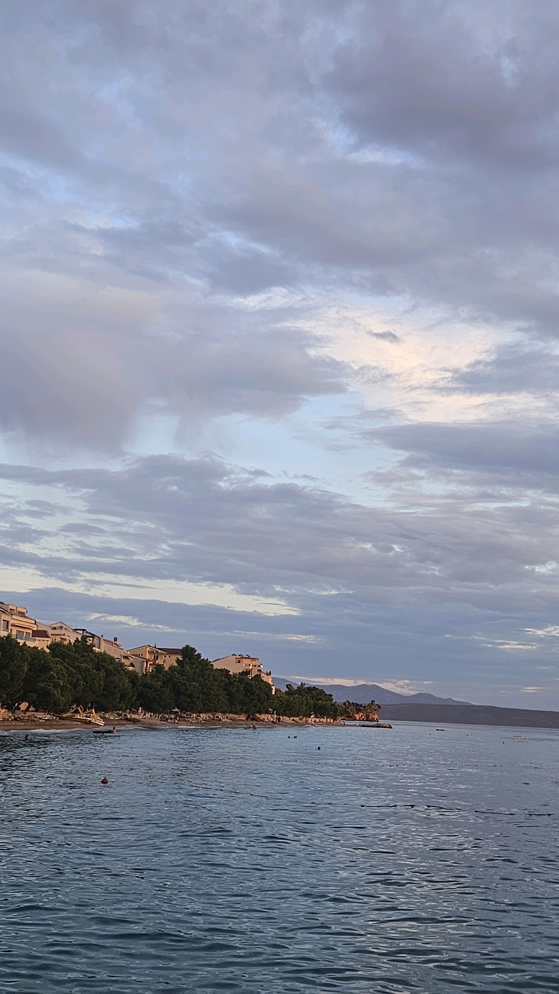 The image depicts a serene coastal scene at dusk in Općina Tučepi, Croatia. The sky is filled with a dramatic display of clouds, painted in hues of blue, grey, and hints of pink and white, suggesting the last light of day. Below the sky, a line of white buildings with terracotta roofs is nestled amongst lush green trees, perched on a gentle slope overlooking the sea. The foreground is dominated by the calm, rippling surface of the Adriatic Sea, reflecting the muted colors of the sky. Scattered across the water are a few buoys marking a swimming area, and several small, dark shapes indicate people swimming or wading. A narrow beach stretches along the coastline, dotted with small figures enjoying the evening. In the distance, a chain of hazy, blue mountains forms the horizon. The overall atmosphere is tranquil and picturesque, capturing the peaceful transition from day to night in a Mediterranean coastal town.