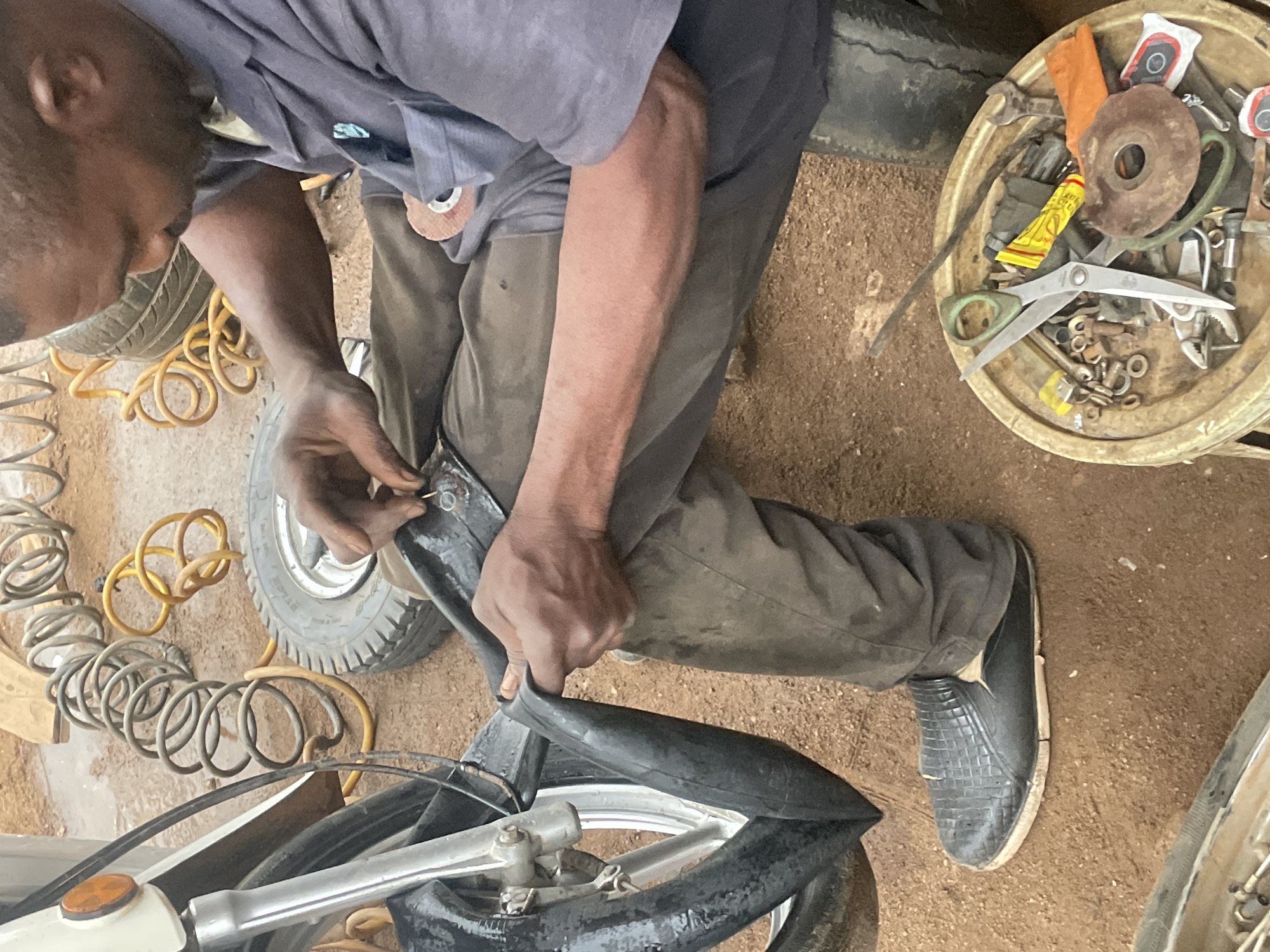 A high-angle shot captures a man working on a motorcycle or bicycle tire in an outdoor repair setting in Jos, Nigeria. The man, with dark skin and short dark hair, is seen from above, wearing a grey short-sleeved top and dark trousers. He is crouched down, intently focused on manipulating a deflated black inner tube or tire with both hands, likely in the process of patching or removing it from the wheel rim.
Around him on the reddish-brown, sandy ground are various tools and components. To his left, a grey coiled air hose and a yellow air hose are visible, along with another small, worn tire. To his right, a circular, light-colored basin is filled with an assortment of hardware, including nuts, bolts, washers, a wrench, a pair of green-handled scissors, a rusty grinding disc, and a yellow tube that appears to be glue or sealant. Parts of the vehicle's frame, front wheel, and handlebars are visible in the lower left, confirming it is a two-wheeled vehicle. The man wears a black, textured shoe on his right foot. The scene is illuminated by natural light, indicating daytime. The environment suggests an informal or roadside repair shop, characteristic of practical, hands-on mechanical work.