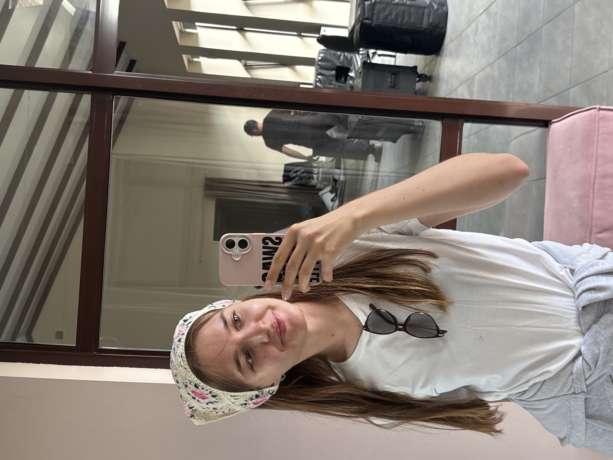 A young woman with long brown hair and a patterned headscarf is taking a selfie in a room with a mirrored wall and a large window. She is wearing a white t-shirt and grey sweatpants tied around her waist. A pair of sunglasses hangs from the neckline of her t-shirt. In the reflection, a man in a black t-shirt and pants is visible, standing near some equipment, possibly in a studio or rehearsal space. The room appears to be sparsely furnished with chairs and cases. The flooring is tiled. The overall impression is of a casual, behind-the-scenes setting, not specific to any particular city.