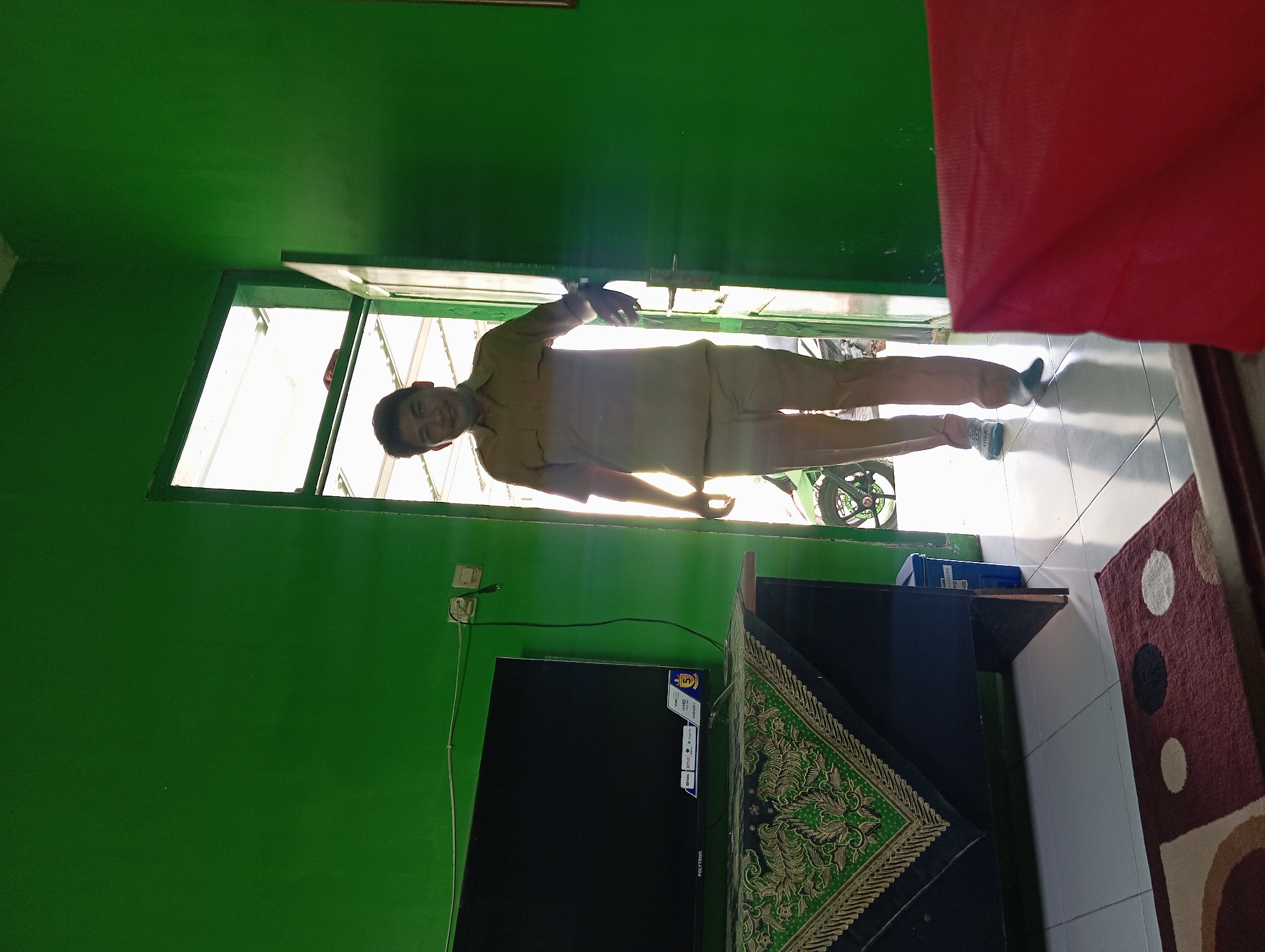 The image depicts an indoor scene with a man standing in a doorway. The walls are painted a vibrant green. To the left, a black flat-screen television is placed on a dark wooden table, which is draped with a patterned green and gold cloth. An electrical outlet and switch are visible on the green wall above the television. The man, wearing a tan collared shirt and matching pants, is walking through the doorway, illuminated by sunlight from outside. He appears to be smiling and looking towards the camera. Through the open doorway, a motorcycle is partially visible outside. In the foreground, a red object, possibly a piece of furniture or fabric, is in focus. A maroon rug with circular patterns is on the floor in the lower left corner. The flooring appears to be white tiles. The scene suggests a residential or office interior in Tegalciut, Indonesia.