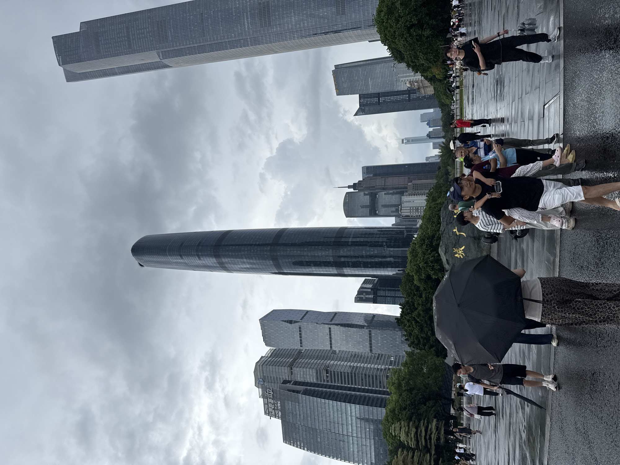 In Tianhe District, China, a group of people are gathered on a wet plaza with numerous skyscrapers in the background. A man in a black t-shirt and white shorts, with a child on his shoulders, walks forward on the plaza. To his left, a person holding an open black umbrella and another person with a closed umbrella stand near each other. Several other individuals are dispersed across the plaza, some walking and others standing. The ground appears wet, indicating recent rain, and the sky is overcast. The architecture of the skyscrapers is modern and varied, with glass facades reflecting the cloudy sky. A large stone with golden Chinese characters is visible on the plaza.