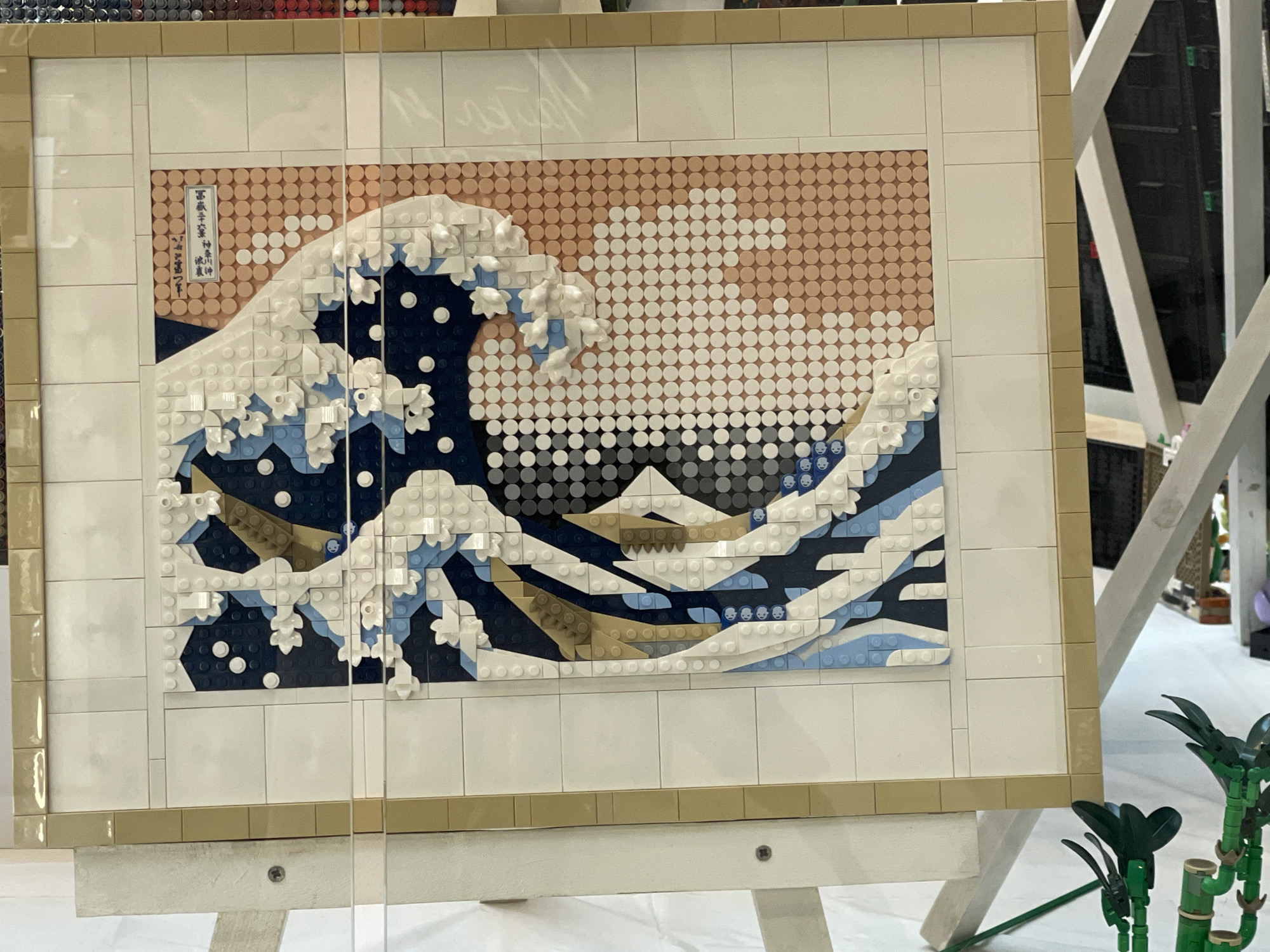 The image shows a detailed Lego mosaic of Hokusai's "The Great Wave off Kanagawa," displayed on an easel. The mosaic is framed with beige Lego bricks and is presented behind a clear protective barrier, indicated by reflections on the surface. The artwork itself is meticulously constructed using a variety of Lego studs in different colors to represent the iconic wave, Mount Fuji in the background, and a Japanese inscription in the upper left corner. The easel supporting the mosaic is white and made of Lego bricks, with visible screws. In the background, out of focus, are other Lego creations and structures, including what appear to be display stands and possibly other models. The scene suggests an exhibition or convention setting, but the specific location in Sofia, Bulgaria, cannot be determined from the visual information provided. There are no people directly interacting with the mosaic.
