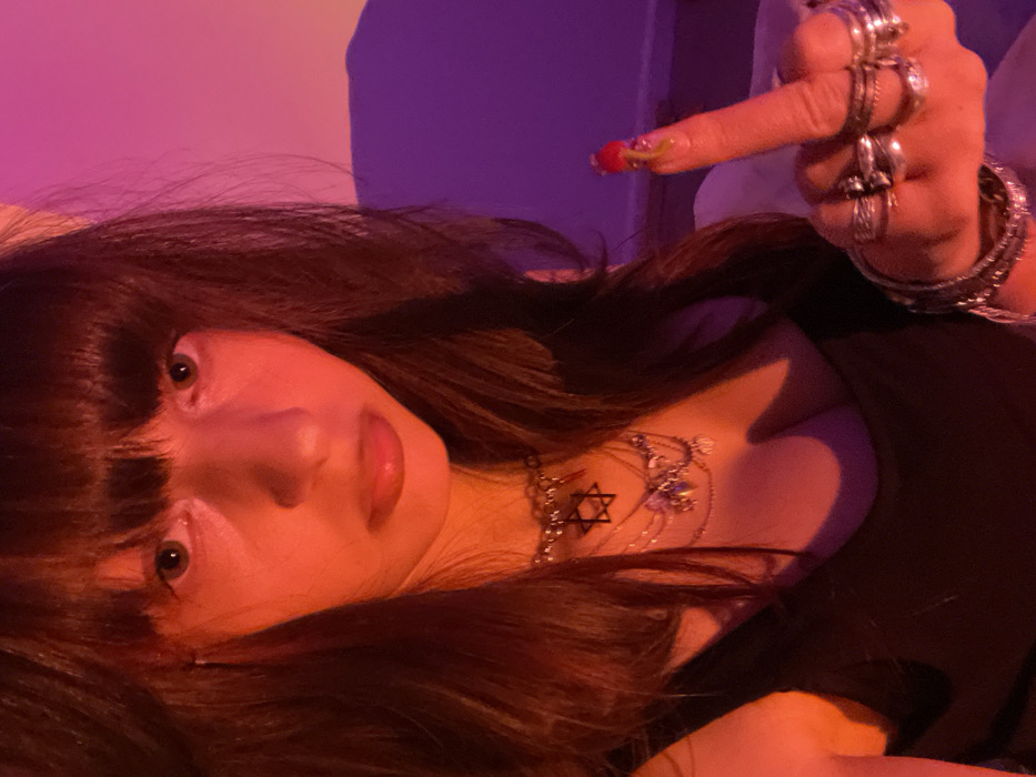 A woman with dark hair and bangs stares intensely into the camera. She wears a necklace featuring a Star of David. Her hand is raised, flipping off the viewer, adorned with numerous rings and bracelets. The scene is lit with a red and purple hue, highlighting her features and jewelry. Her nails are painted with a unique, colorful design. She wears a black top that reveals cleavage.