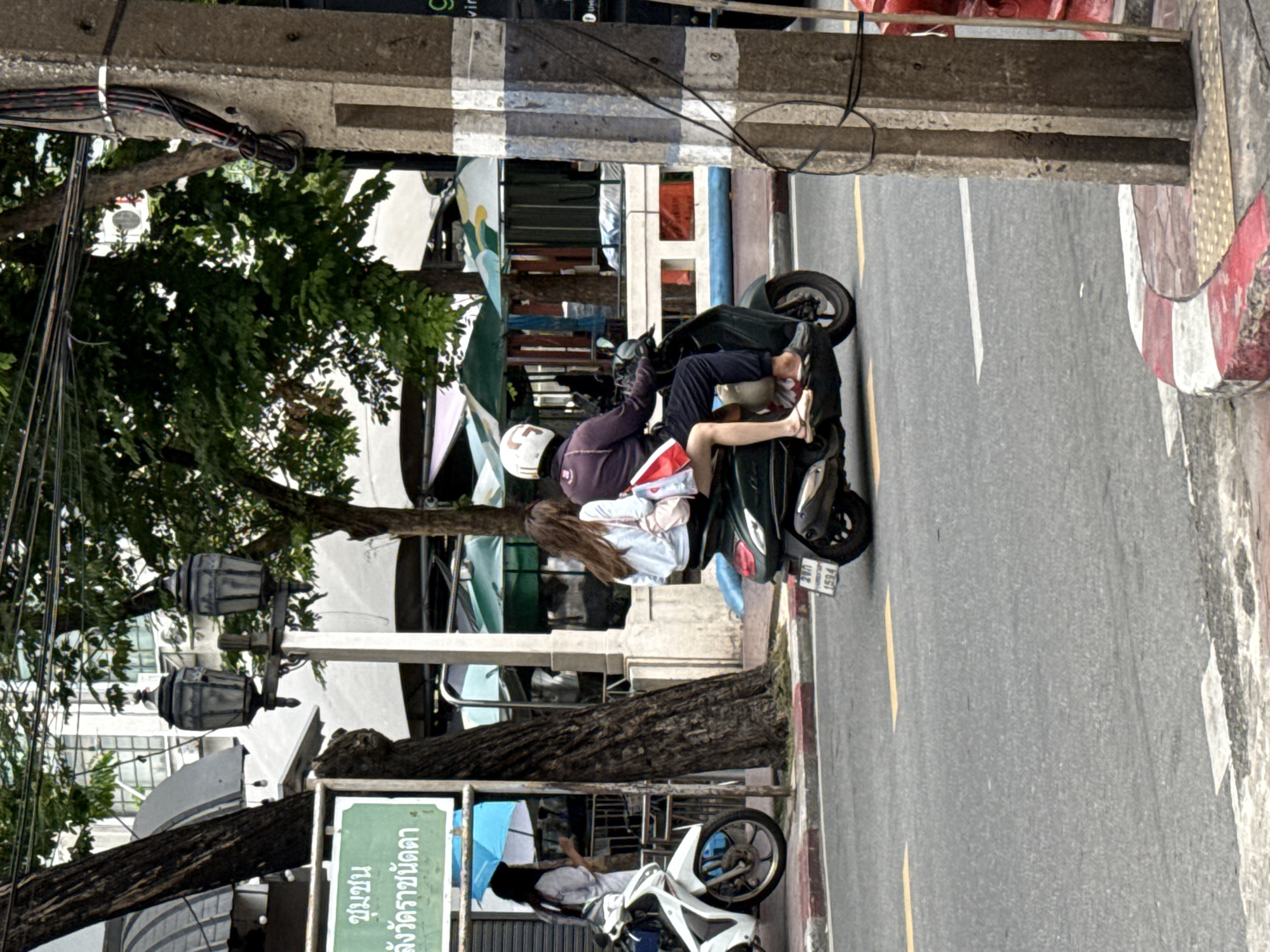 In Bangkok, Thailand, on a street with a paved road and sidewalk, a dark-colored scooter travels away from the viewer. The scooter is occupied by two individuals. The person in the rear, wearing a light blue long-sleeved shirt and dark pants, has long brown hair and is carrying a pink bag and red and white bags. The person in the front, wearing a dark purple long-sleeved jacket and dark pants, has a white helmet with black markings. The scooter has a license plate with the characters "280" and "1594". To the left of the scooter, a white motorcycle is parked. Behind the scooter, a light-colored utility pole with wires is visible, along with trees and buildings. A green sign with Thai text is mounted on a metal pole in the background.