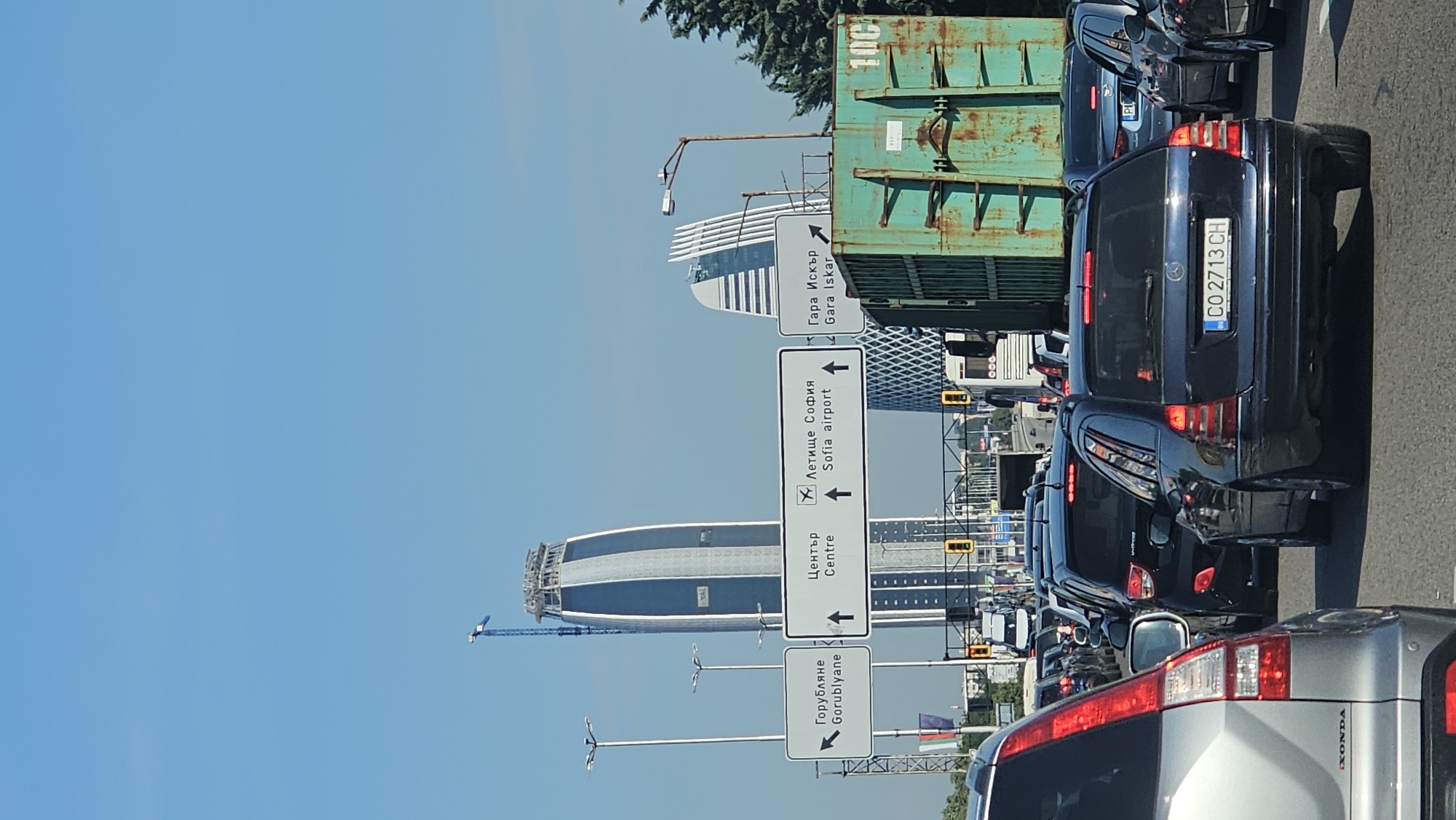 The image captures a traffic jam in Sofia, Bulgaria, with a clear blue sky overhead. In the foreground, several cars are stationary on a road. A silver SUV is on the left, and a dark blue Mercedes-Benz wagon is prominently in the center, displaying a license plate reading "CO 2713 CH". Behind it, other vehicles are visible.
Dominating the background are modern skyscrapers, one of which appears to be under construction with a crane at its apex. Road signs in Bulgarian and English indicate directions to "Gorublyane", "Center", "Sofia airport", and "Gara Iskar". A large, green, rusted shipping container is positioned to the right of the road, adding to the industrial feel of the scene. Traffic lights are visible on an overhead gantry, indicating a stop or slow down. The overall impression is one of a busy urban environment experiencing a traffic delay.