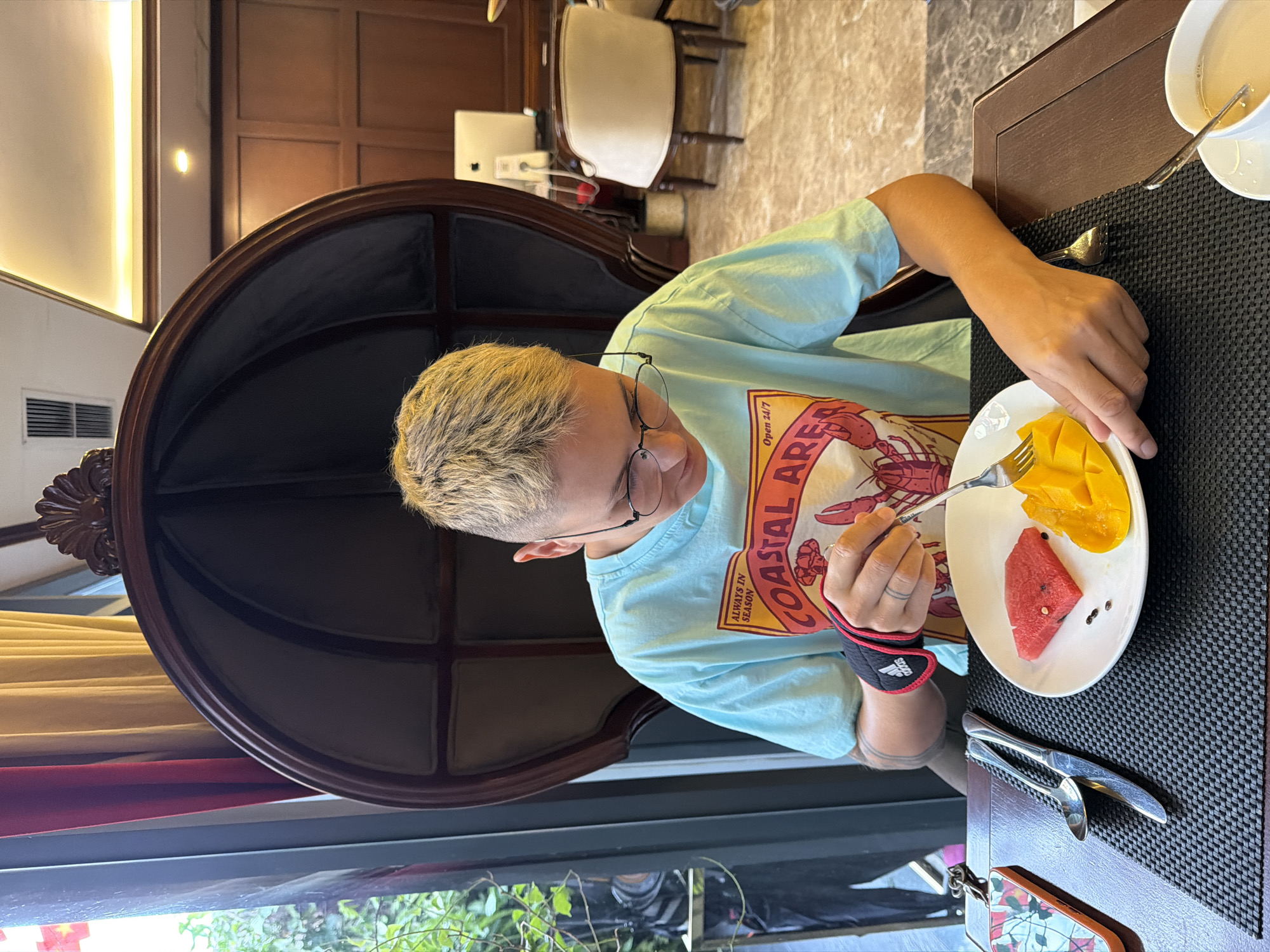 A person with short, dyed blonde hair and glasses is seated in a large, dark upholstered chair and eating fruit from a plate. The person is wearing a light blue t-shirt with a graphic of a lobster and text that reads "Coastal Area". A black wristband with red trim and the Adidas logo is on the person's left wrist. On the plate are two slices of watermelon and a portion of sliced mango. The person is using a fork to pick up a piece of mango. The scene appears to be indoors, possibly a hotel lobby or restaurant, with warm lighting. In the background, there are curtains, wooden paneling, and office furniture. The location is Thuận Hóa Ward, Vietnam.