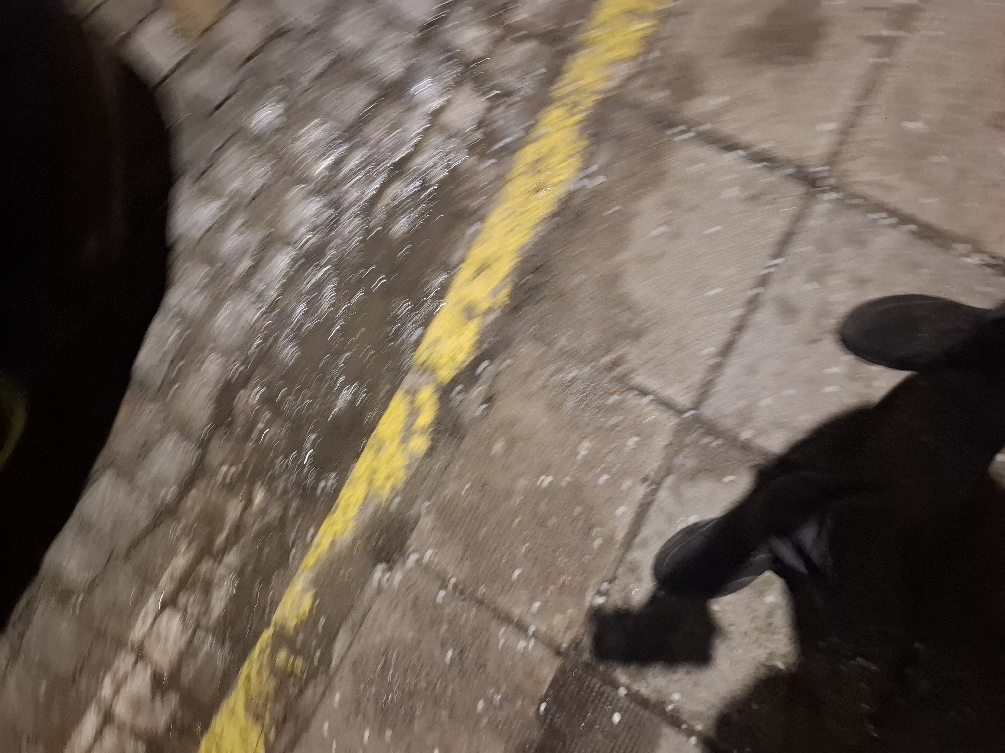 The image depicts a poorly lit sidewalk in Sofia, Bulgaria. A yellow line marks the edge of a walkway, adjacent to what appears to be a low wall. The sidewalk is made of paving stones. Debris, possibly small pieces of snow, is scattered across the surface. The shadow of a person's legs and feet is visible, suggesting a first-person perspective.