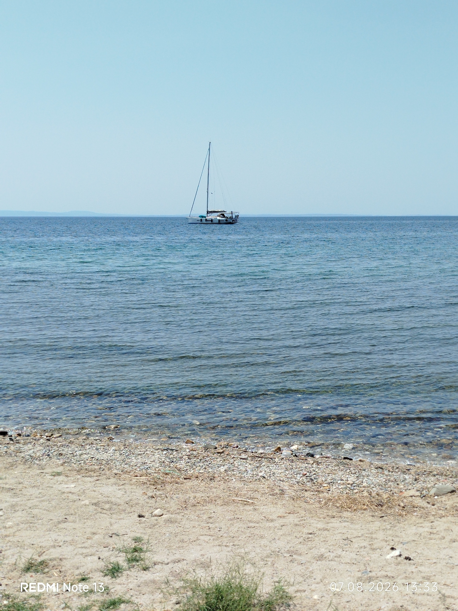 A sailboat is anchored in the clear, calm water off a pebble and sand beach. The sky is a pale blue, and a hazy landmass is visible on the horizon. The image is taken from the beach, looking out towards the water. The bottom left corner displays "REDMI Note 13", and the bottom right corner shows "07.08.2026 13:33". This scene is typical of the coastal areas within the Sithonia Municipal Unit in Greece, known for its tranquil waters and picturesque beaches.