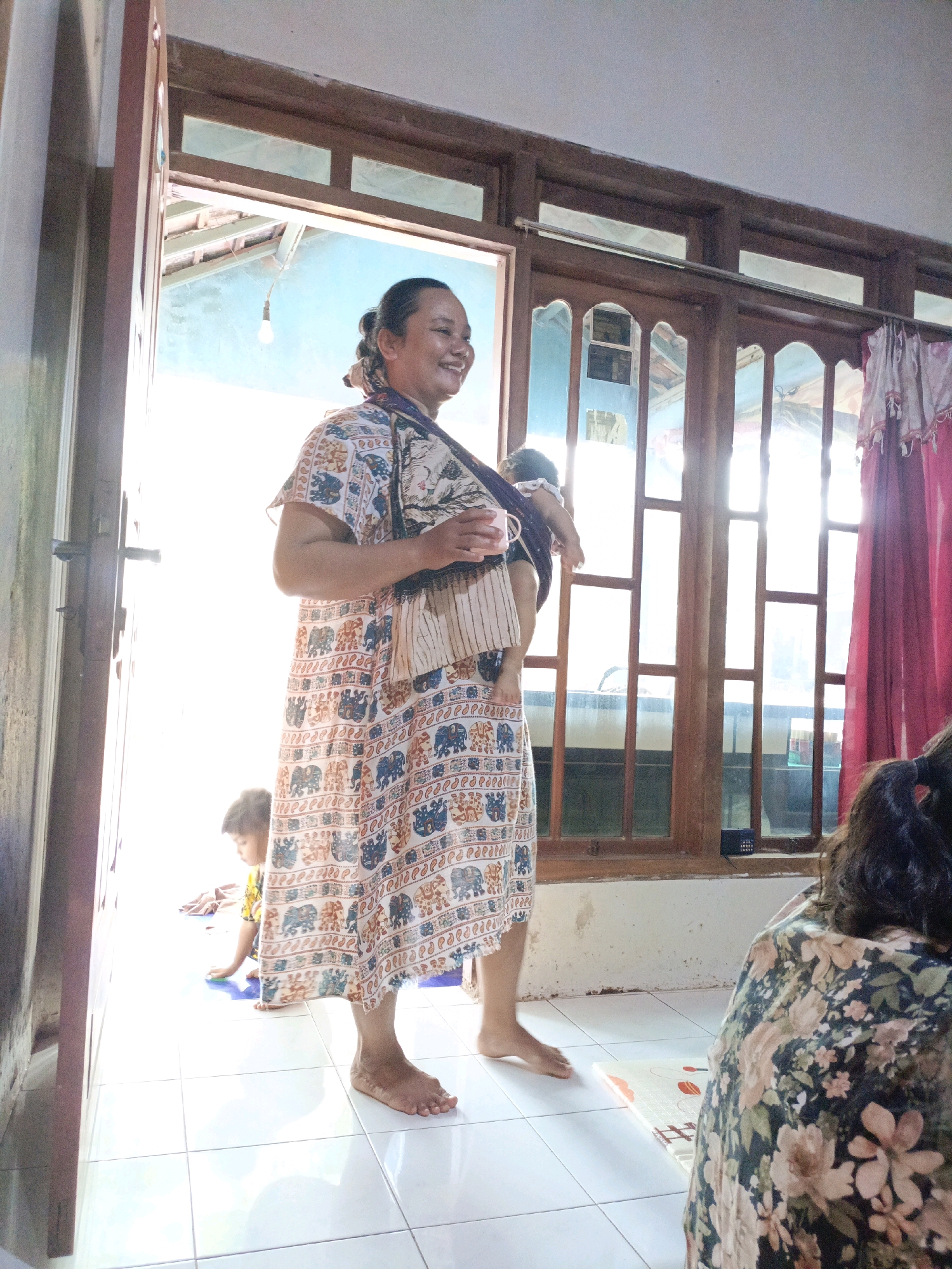 A woman carrying a baby on her back stands in a doorway, smiling and looking to her right. The baby, partially visible, is wrapped in a fabric sling. The woman is wearing a long, patterned dress with elephants and floral motifs. Sunlight streams in from behind her, creating a bright background. To her left, a young child in a yellow shirt sits on the floor. To her right, a woman with dark hair is seated, facing away from the camera, wearing a floral patterned top. The scene appears to be indoors, with tiled flooring and wooden window frames visible. This image was captured in Jenu, Indonesia.