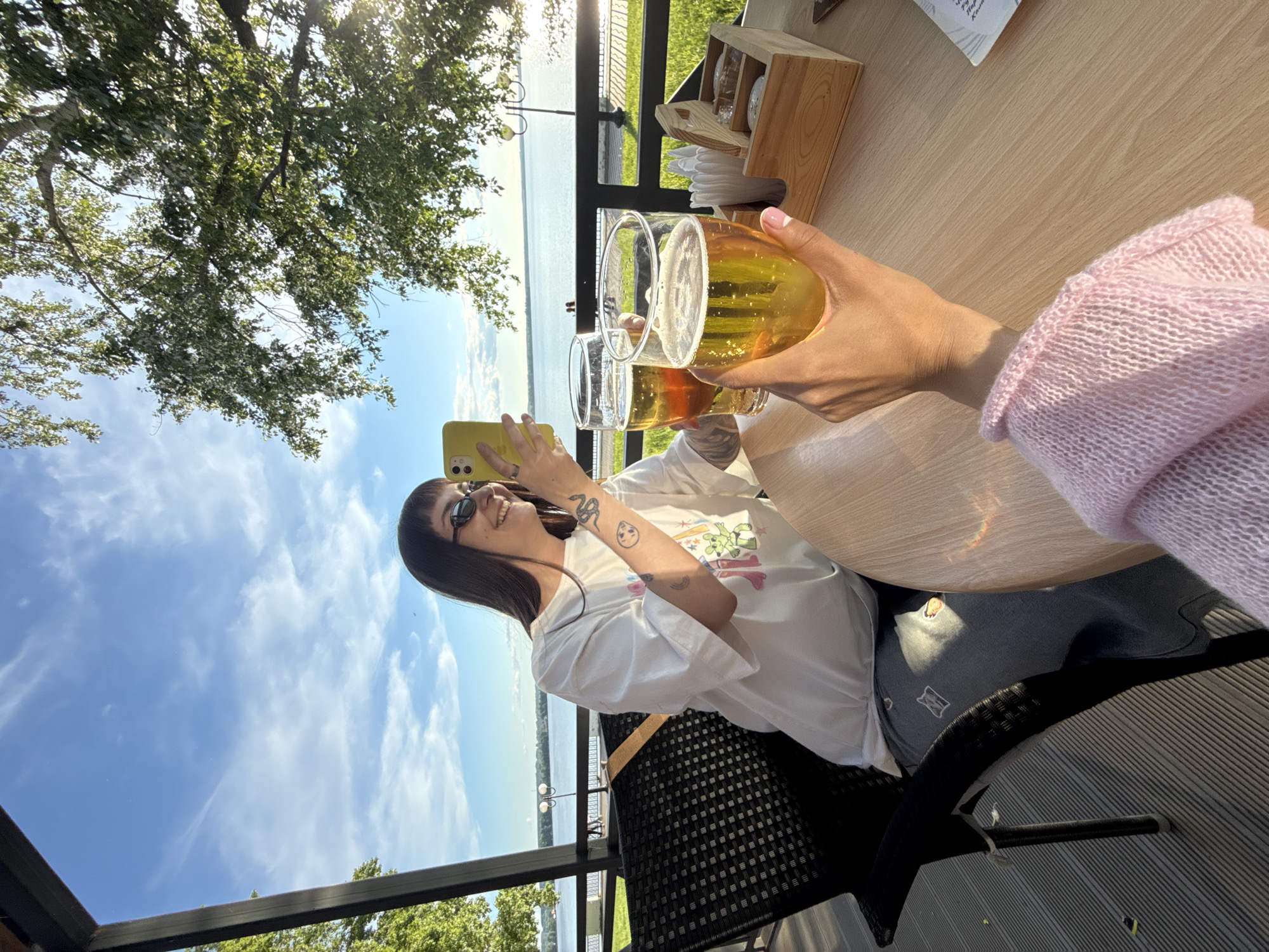The image shows a cheerful outdoor scene at a restaurant or cafe overlooking a large body of water, likely in Votkinsk, Russia. The perspective is from a table, looking out towards the horizon.
Two individuals are partially visible. In the immediate foreground, the hand and arm of one person, wearing a light pink knitted sweater, hold two tall glasses filled with a golden, foamy beverage, likely beer or a similar drink, clinking them together as if making a toast. Opposite, a woman with dark hair and bangs, wearing dark sunglasses and a white t-shirt with colorful cartoon-like graphics, is smiling broadly. She holds a bright yellow smartphone up, seemingly capturing a moment or taking a photo. Tattoos, including a snake design and a round graphic, are visible on her arm. She is seated in a dark, patterned outdoor chair.
The setting features a light wooden table, on which a wooden caddy with napkins and other items is placed. Beyond the table, a black metal railing separates the dining area from an expansive body of water, possibly the Votkinsk Reservoir. A distant shoreline with trees and scattered buildings or structures is visible across the water. The sky is clear and blue with some scattered white clouds. Bright sunshine illuminates the scene, creating a warm glow and some lens flare, suggesting a sunny daytime, possibly late afternoon. Lush green tree foliage is prominent in the upper left corner of the frame. The overall atmosphere conveyed is one of relaxation and enjoyment. No discernible text is visible in the image.