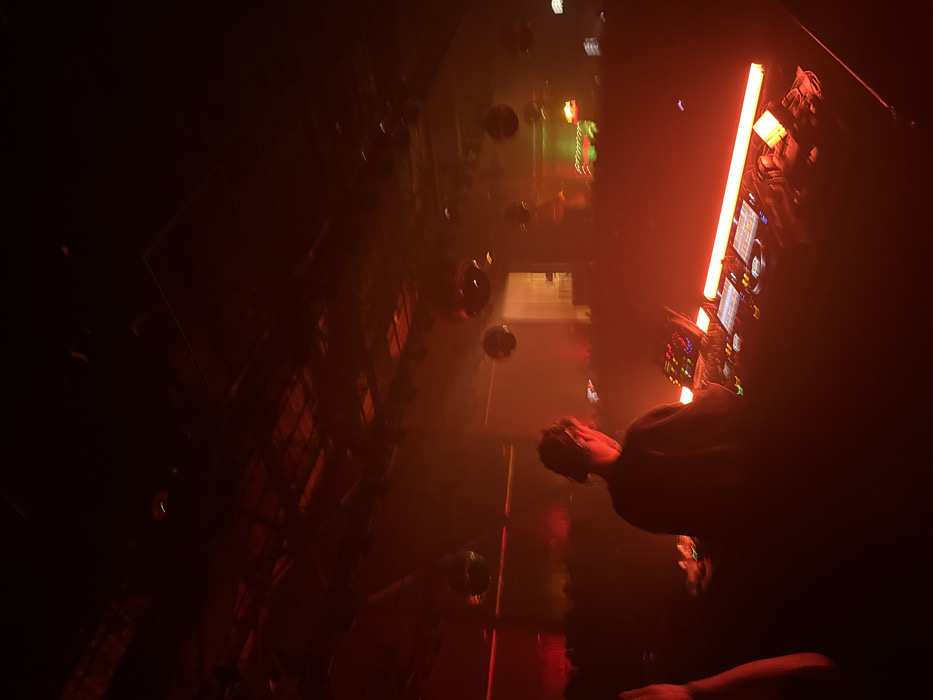 The image depicts an indoor scene at an electronic music venue, specifically identified as Sound Nightclub, located at 1642 Las Palmas Ave, Hollywood, Los Angeles, California, United States of America.
The environment is characterized by low light, heavy atmospheric haze, and a dominant red illumination. An individual, presumably a DJ, is positioned centrally in the foreground, viewed from behind, wearing a cap and a dark top. This individual is actively engaged with a professional DJ setup, which includes at least two media players and a central mixer, all emitting subtle operational lights. A prominent, elongated red LED light bar is situated directly in front of the equipment, brightly illuminating the DJ's hands and the console. A smaller, square red light source is visible to the right of the equipment.
The background reveals a large, dark, open space with an indistinct crowd of individuals visible beyond the DJ booth, partially obscured by the haze and ambient lighting. Multiple reflective disco balls are suspended from the ceiling, distributed across the mid-ground and background. The ceiling itself appears to be an exposed industrial structure with visible beams and utilities. Distant background lighting includes additional red and some green illumination, indicating other stage elements or venue sections. A partial arm and shoulder of another person are visible on the far left edge of the frame, indicating proximity to the photographer.