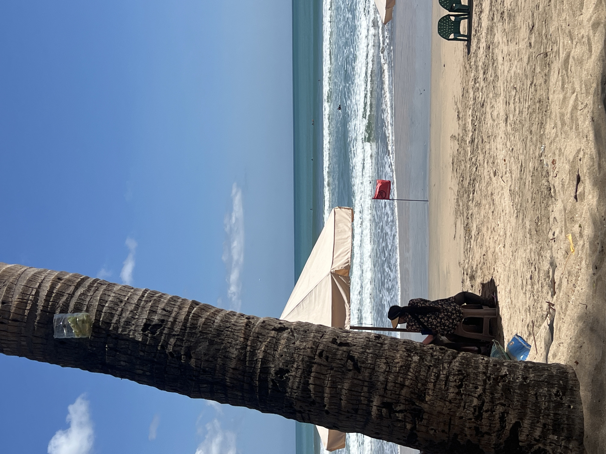 An outdoor shot shows a beach scene in Kuta, Indonesia, on a clear, sunny day. In the foreground, the textured trunk of a palm tree dominates the left side of the frame, with a plastic bottle filled with what appears to be green matter attached to it. Partially visible under a beige beach umbrella, a person wearing a dark, floral-patterned shirt and a hat is seated on a stool, facing away from the camera. Nearby, a blue bag and a green cushion lie on the sand.
The middle ground reveals a sandy beach leading to the ocean. A red flag on a pole stands planted in the sand. In the distance, several surfers are visible in the turquoise water, some riding waves. Two empty green plastic chairs are positioned on the sand to the right. The background is a expanse of clear blue sky with a few scattered white clouds. The overall atmosphere is serene and tropical.
