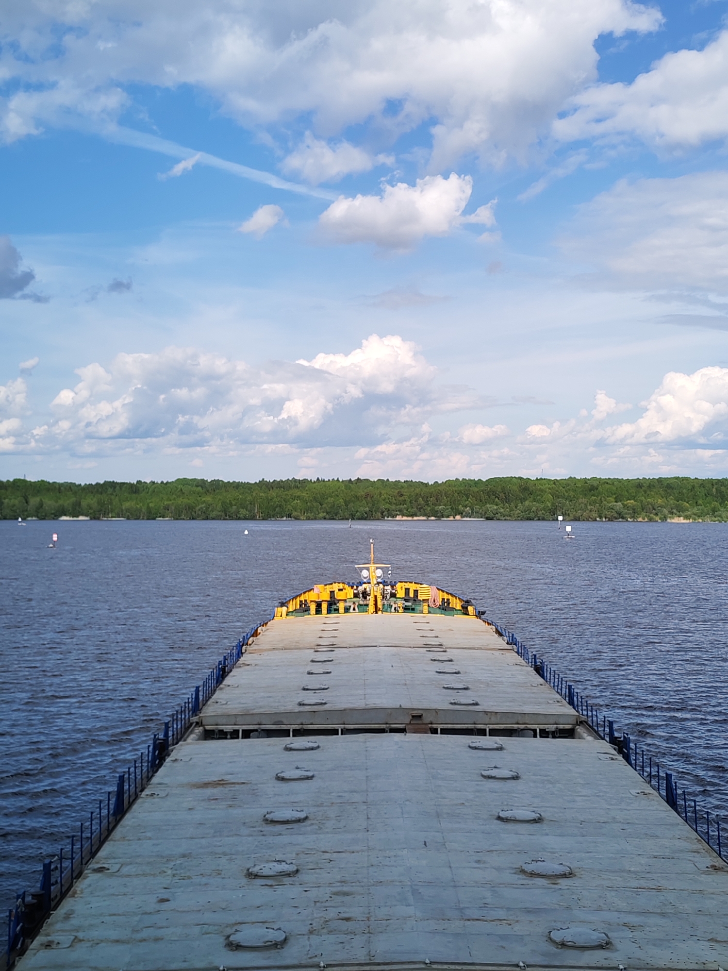 The image shows the deck of a barge traveling on a body of water, presumably a river or lake, with a forested shoreline in the distance. The sky is blue with scattered cumulus clouds.
The deck of the barge is a gray, flat surface with rows of circular hatches. Blue railings line the sides of the deck. Towards the far end of the barge, a yellow structure with various equipment is visible.
The water is calm with slight ripples, reflecting the blue of the sky. The distant shoreline is densely forested.
The location is Novodevich'e, Russia. This information is based on common knowledge of barge transportation on Russian waterways.