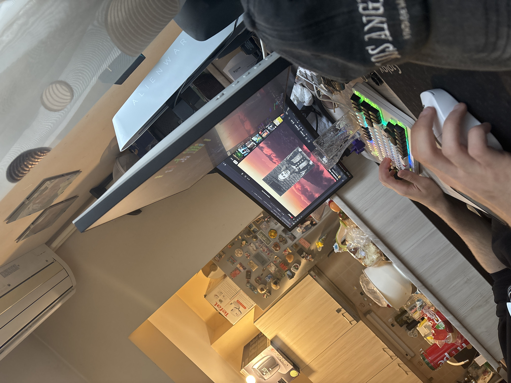A person wearing a dark-colored "Los Angeles" baseball cap is using a white computer mouse on a desk with a multi-colored keyboard. An Alienware computer tower sits behind a computer monitor displaying a photograph of a person. A glass cup rests on the desk near the keyboard. The desk is situated in a kitchen, where cabinets, an air conditioning unit, and various kitchen appliances, including a Tefal product, are visible. The location is not definitively identifiable as Plovdiv, Bulgaria, without more specific contextual clues within the image.
