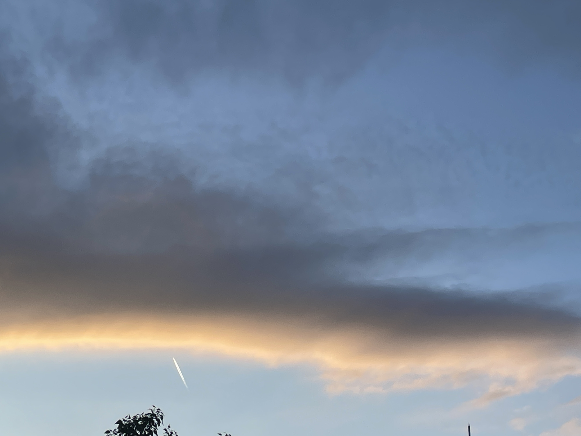 The image shows a sky with dark, ominous clouds. A band of orange light illuminates the bottom edge of the clouds. An airplane contrail is visible in the lower left of the image. In the bottom of the image are some leafy tree branches and an object that could be a radio tower or a building.
The location is Unknown, Unknown.