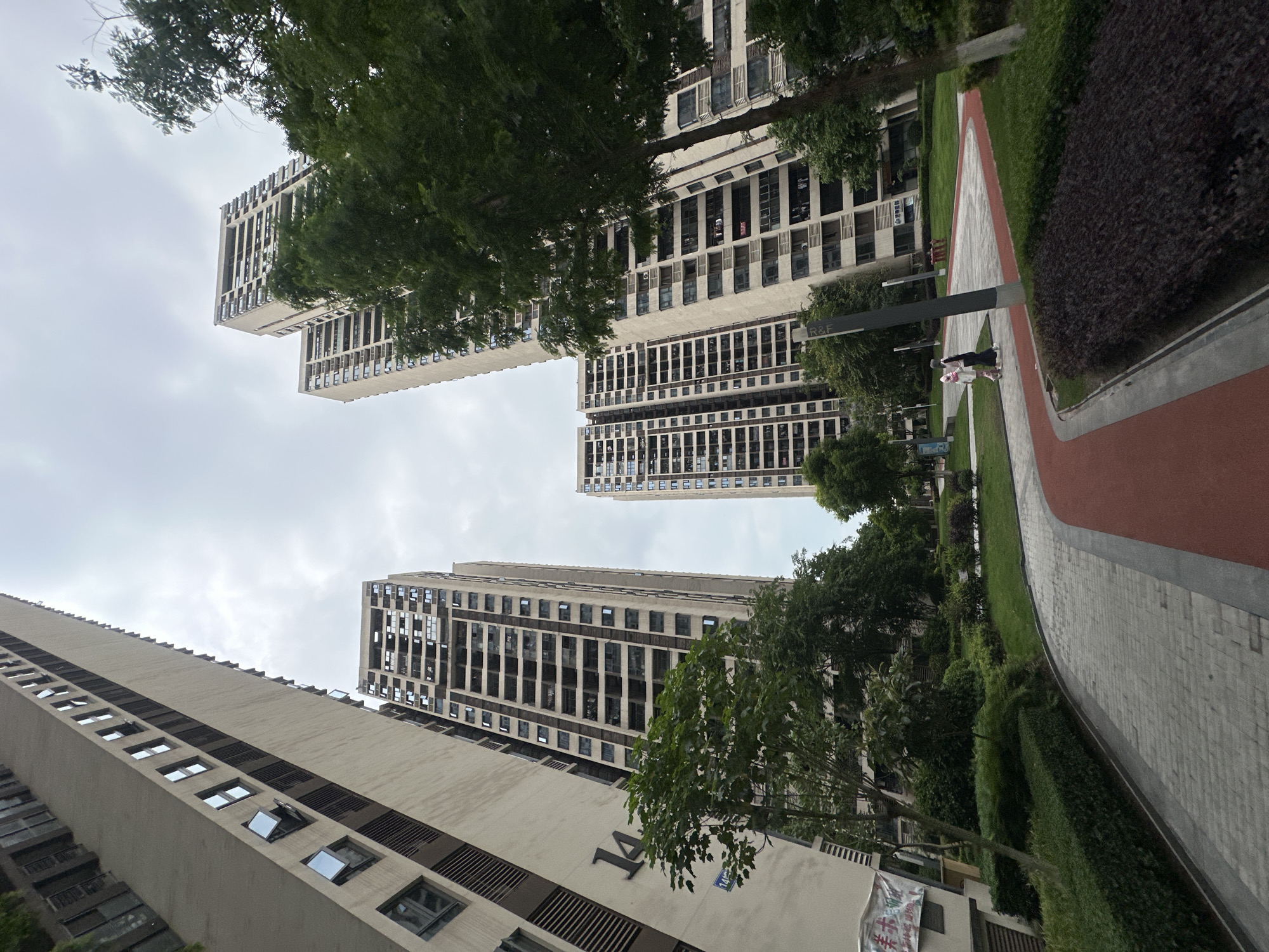 The image, taken from a low angle in Chongqing High-tech Zone, China, showcases a cluster of modern high-rise buildings under a cloudy sky. The buildings are characterized by their multiple rows of windows and neutral color scheme. Trees and green foliage partially obscure the lower sections of the buildings. A paved pathway with a red running track runs alongside a patch of green grass. There are two people walking on the pathway and there is a road sign that is partially visible.