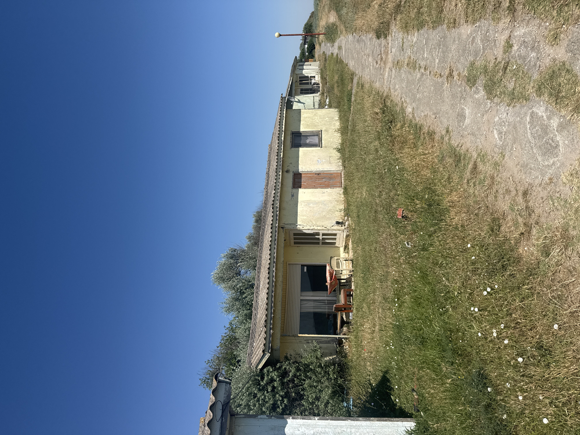 The image depicts a sunny day in Shabla, Bulgaria, showing a row of small, weathered bungalows lining a dirt path. The bungalows, painted yellow and with corrugated roofs, appear somewhat neglected. In front of one bungalow, a small table with chairs sits in a grassy area dotted with wildflowers. The path leads towards a light pole and the distant horizon, suggesting a coastal or open landscape. The overall scene conveys a sense of quiet abandonment or a rustic, seaside retreat.