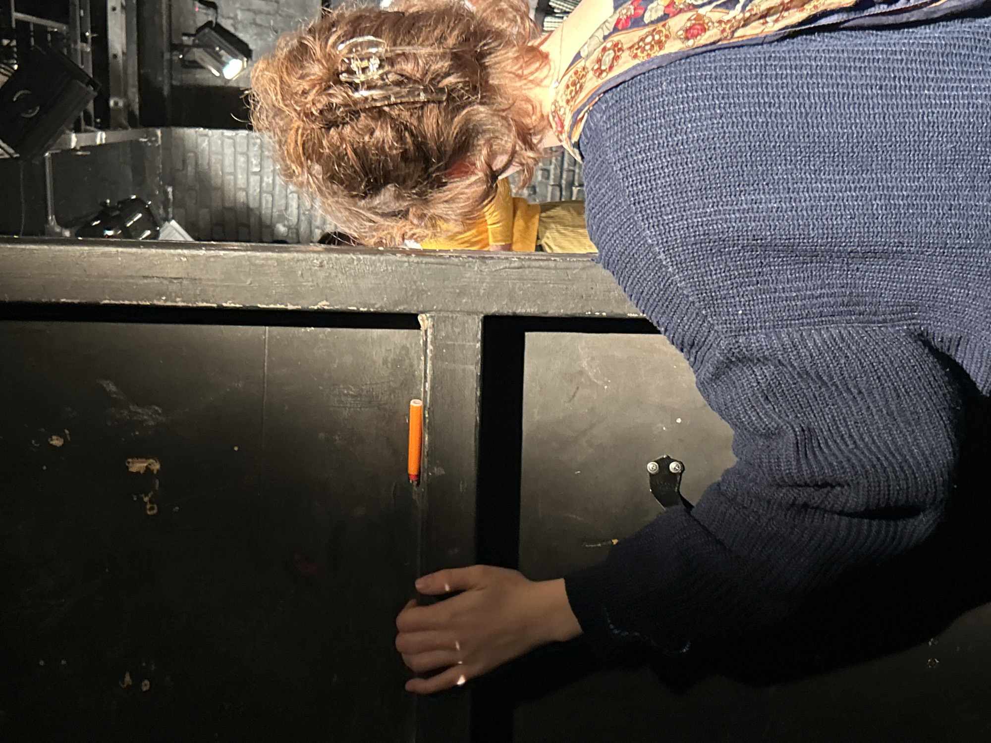 A person with light skin, wearing a dark blue knitted sweater and a patterned scarf around their neck, is seen partially in the frame, bending over and reaching into a dark, likely wooden, cabinet or storage unit. Their hand is visible near the door of the cabinet, which has an orange marker stuck between the doors. The background is indistinct, but what is visible suggests a backstage or utilitarian area, possibly including lighting equipment. The location is not discernible from the given image.