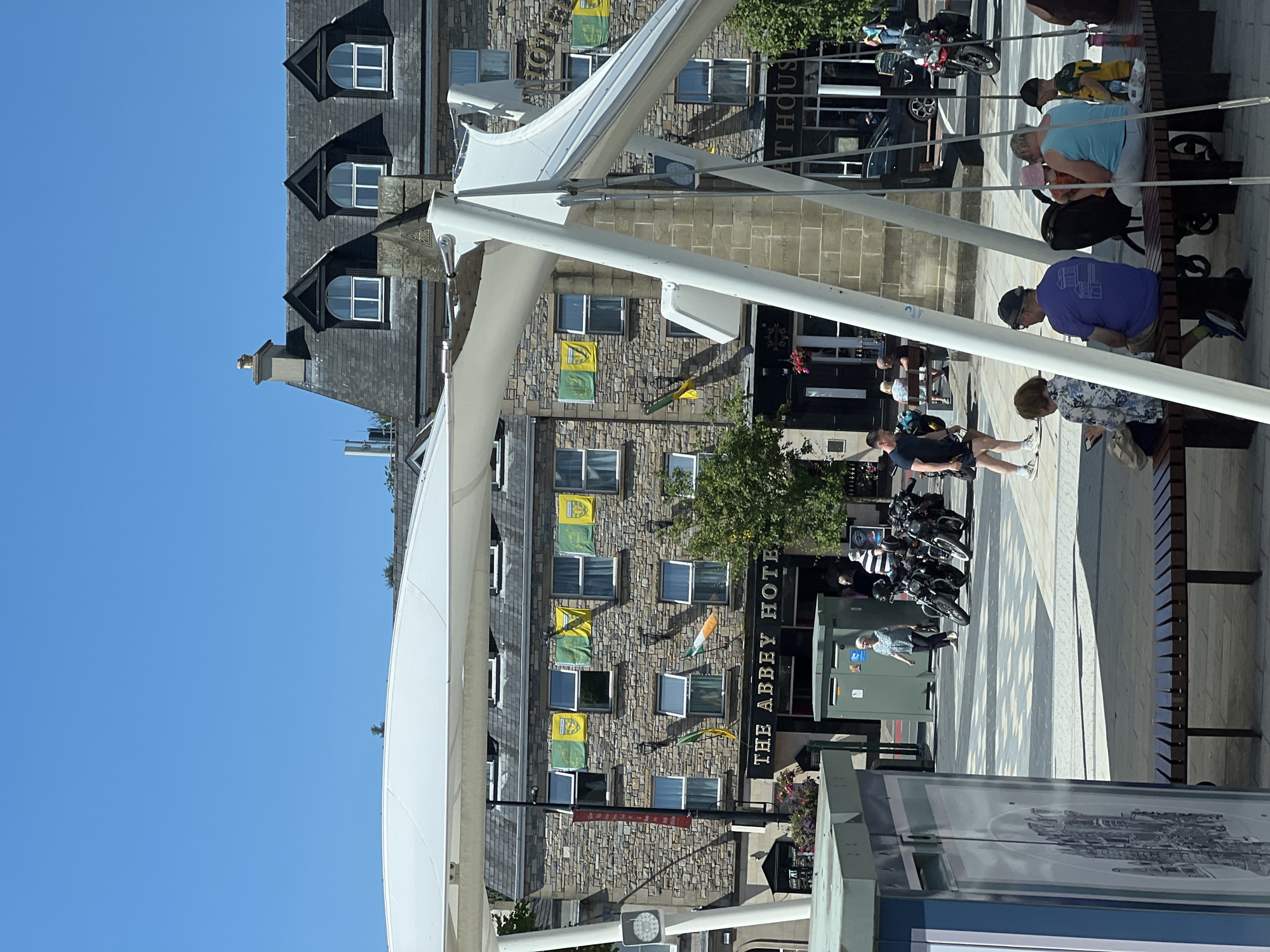 The image depicts a street scene in Donegal Town, Ireland, in front of The Abbey Hotel. A white modern canopy structure dominates the foreground. The Abbey Hotel, a stone-faced building, is visible behind the canopy, adorned with green and yellow flags, some of which appear to be Gaelic football county flags, and an Irish tricolor flag. Several motorcycles are parked in front of the hotel entrance.
In the foreground, a group of people are seated on a wooden bench. A woman in a floral shirt is looking at her phone, a man in a purple t-shirt and a hat is seated next to her. Further down the bench, a woman in a light blue tank top holds a baby in a pink hat, and a child in a green and yellow jersey sits beside her. A stroller is parked behind the people on the bench.
In the middle ground, an elderly woman in a blue patterned shirt walks past a green bin. Other individuals are visible in the background, including a man in a black t-shirt and shorts standing near the motorcycles, and a few people near the entrance of the hotel. The ground is paved with a patterned design of grey and white squares and lines. The sky is a clear, bright blue, indicating a sunny day.
