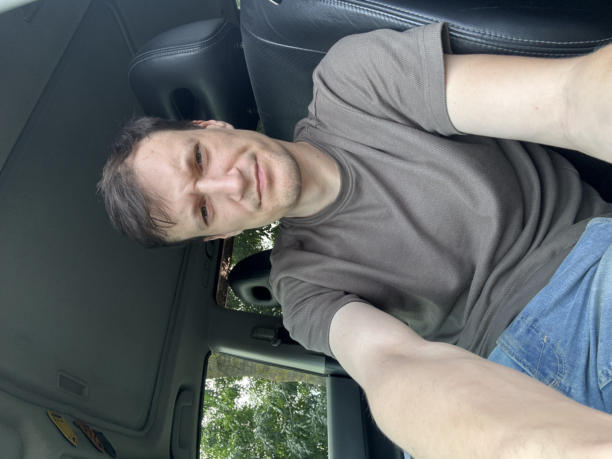 A man is sitting in the driver's seat of a car, looking at the camera with a slightly furrowed brow. He is wearing a brown, textured t-shirt and blue jeans. The car's interior is visible, including the black leather headrest of the passenger seat and the car's sunroof. Outside the car, green trees and foliage can be seen through the windows. There are two yellow and brown stickers on the car's headliner near the sunroof. The lighting suggests it is daytime. The location is identified as Makiv, Ukraine.