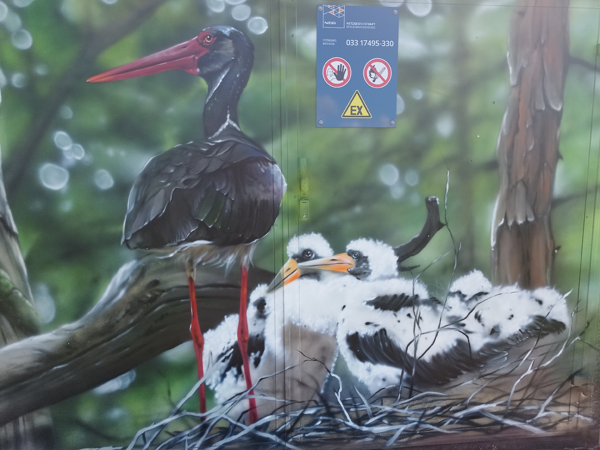 The image shows a painted mural on a metal structure in an unknown city. The mural depicts a black stork standing on a branch next to a nest containing three stork chicks. Above the mural is a blue sign with the logo of NBB Netzgesellschaft Berlin-Brandenburg. The sign includes the phone number 033 17495-330, two warning symbols (one indicating not to touch and another indicating no open flames), and a yellow triangle with the letters "EX" inside. The background is a blurry green with what appears to be tree branches and foliage.