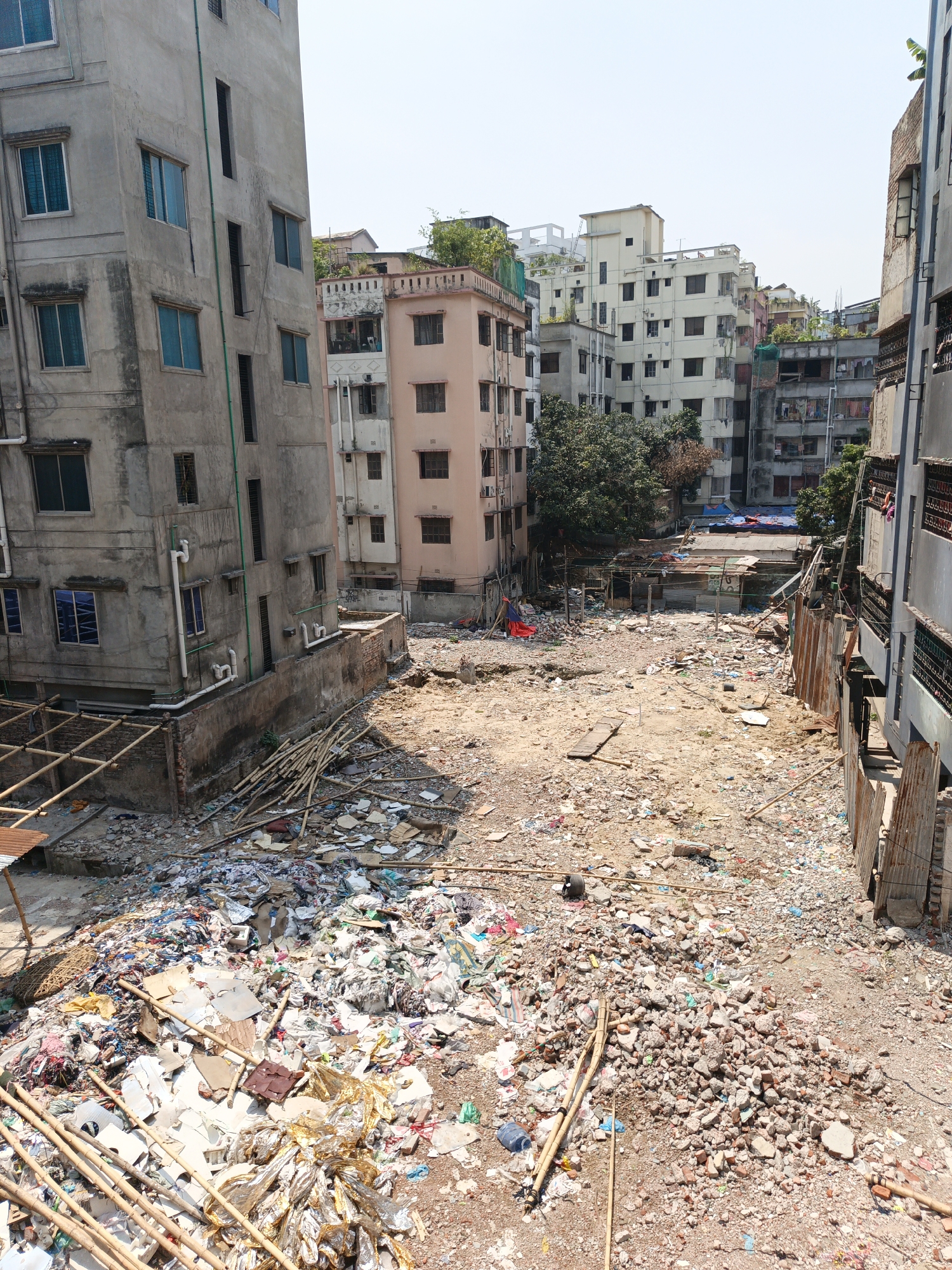 The image shows a vacant lot in Dhaka, Bangladesh, filled with debris and garbage. The lot is surrounded by multi-story buildings, some of which appear to be residential. The ground is covered in a mixture of trash, rubble, and exposed dirt. Bamboo poles are scattered across the foreground, along with discarded construction materials and household waste. In the background, some shacks or temporary structures are visible amidst the refuse. The overall impression is one of urban decay and environmental neglect. There are no people visible in the frame.