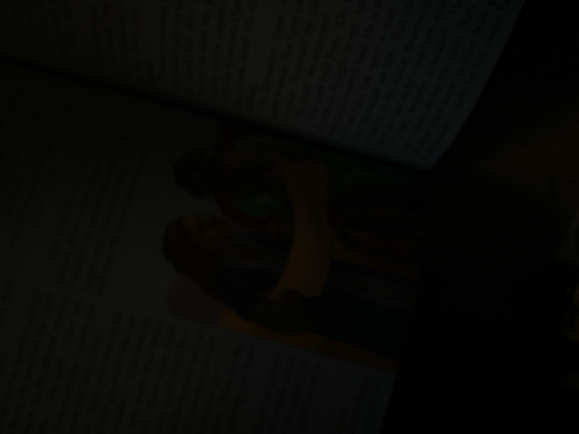 The image shows an open book with text and an illustration. The illustration depicts two figures, possibly from a religious text, with one figure leaning towards the other. The book is angled, and the lighting is dim, casting shadows. The environment is dark, suggesting it might be nighttime or an indoor setting with low light. There is no indication of Jauro Yinu, Nigeria, in the image.