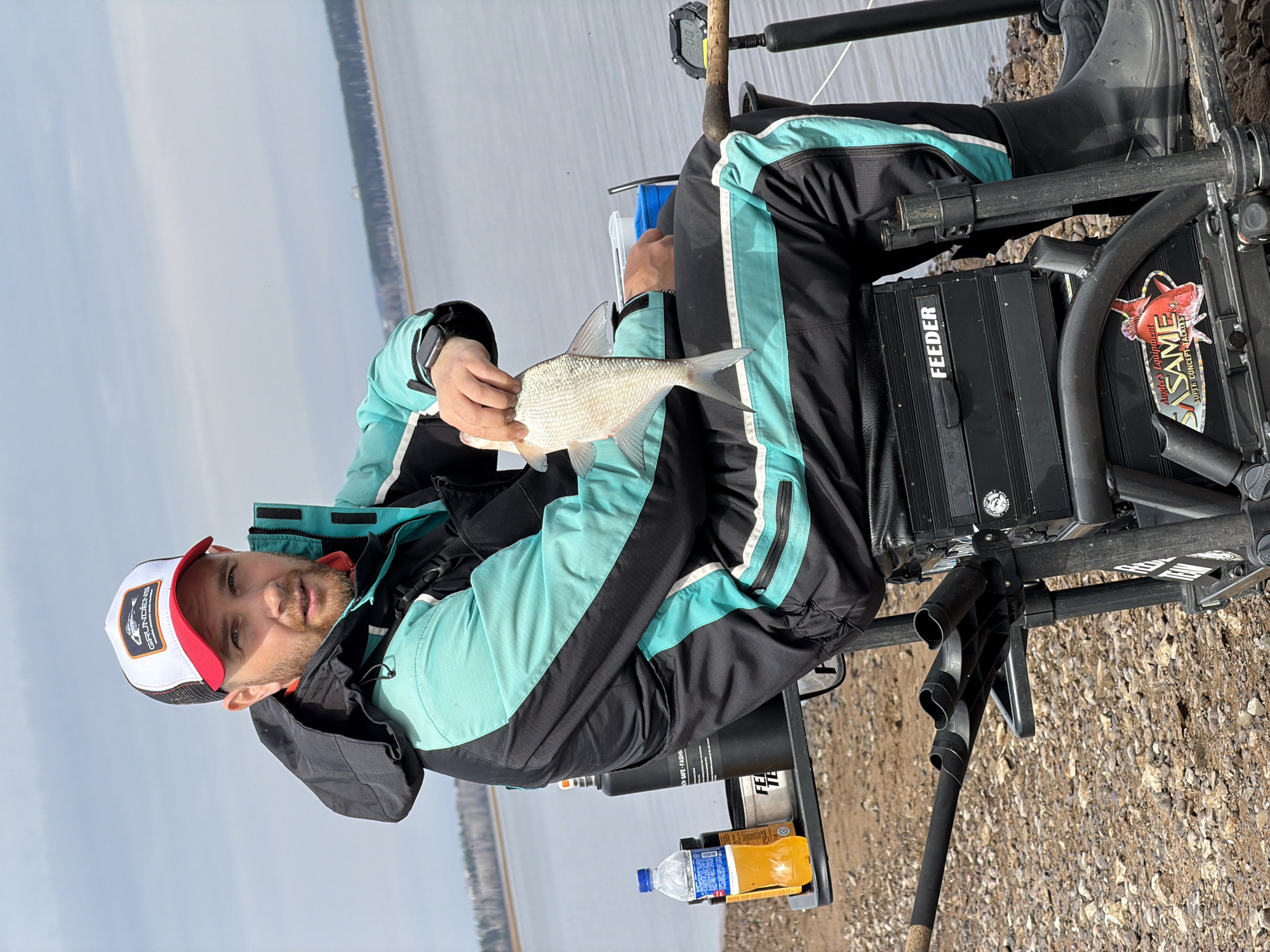 A man in fishing gear sits by a body of water, holding up a fish. He is wearing a black and teal jacket, black pants with teal stripes, and black rubber boots. A white and red hat sits on his head. He is seated in a specialized fishing chair, which has a black tackle box labeled "FEEDER" and a side table holding two bottles. A fishing rod rests on the chair. The background features a wide body of water and a distant treeline. Based on the information provided, this photo was taken in Chaikovsky, Russia.