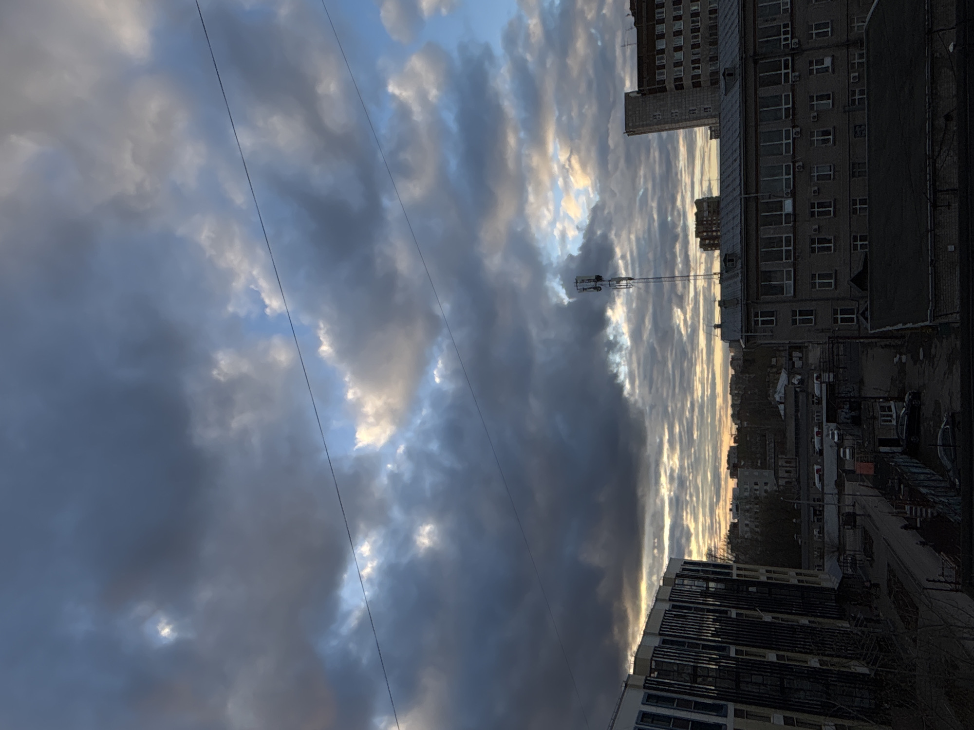 The image depicts an urban scene in Perm, Russia, featuring a view from an elevated position, possibly a window. Dominating the view are heavy, dark clouds filling the sky. These clouds are broken by patches of lighter sky, hinting at a sunset or sunrise.
Several buildings are visible. In the mid-ground, a multi-story building with numerous windows is prominent. There's a telecommunications tower located between the buildings. In the foreground, there's a modern building with visible glass panels and architectural details.
Below, a street scene is present, though details are obscured. Parked cars and hints of foliage are apparent. Overhead, at least two electrical wires stretch across the view.