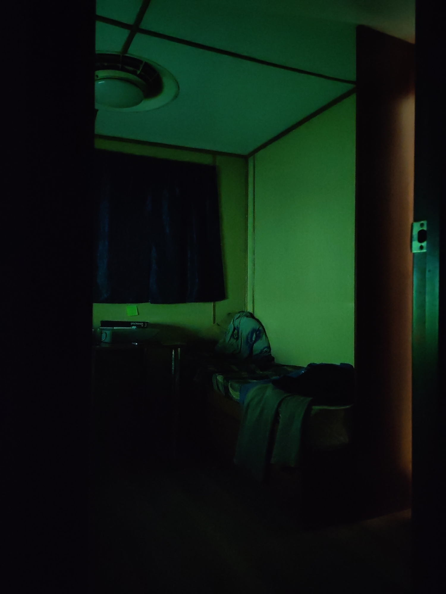 A dimly lit room with green light emanating from an overhead ceiling light. The room contains a bed with rumpled bedding and what appears to be clothing draped over it. Beside the bed is a desk or table with a book and a mug on top. Curtains cover the window. A partially visible doorway is on the right, casting a dark shadow. The location and city are unknown.