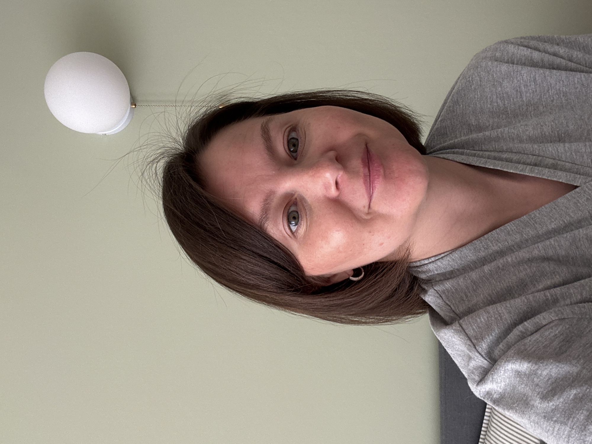 A selfie of a woman with short brown hair, green eyes, and a gray t-shirt. She is looking directly at the camera. A white light fixture is visible in the upper left corner. The background is a light green wall. The location is not discernible from the image.