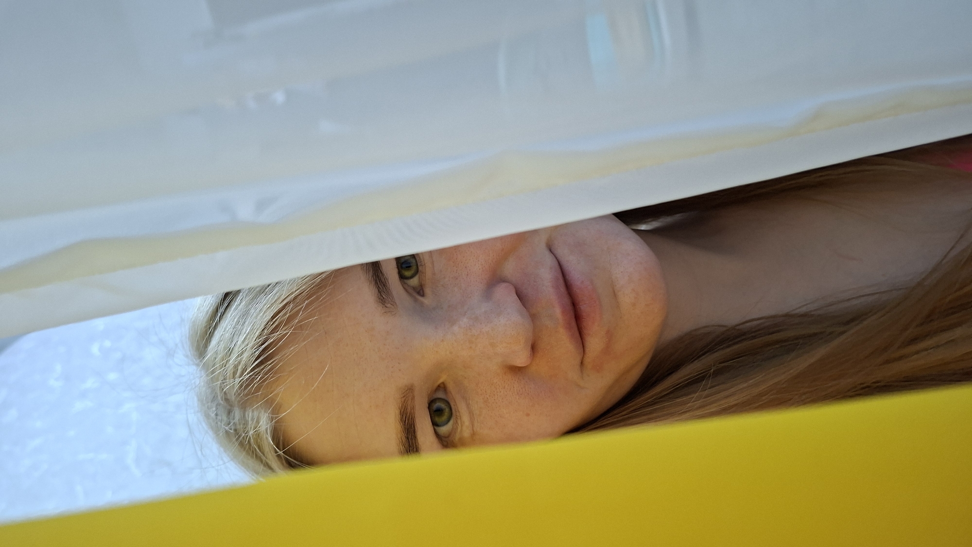 A woman with blonde hair and green eyes is looking through a narrow space between white and yellow surfaces. Her face is visible with freckles. The setting or location within Chaikovsky, Russia, is not discernible from the image.