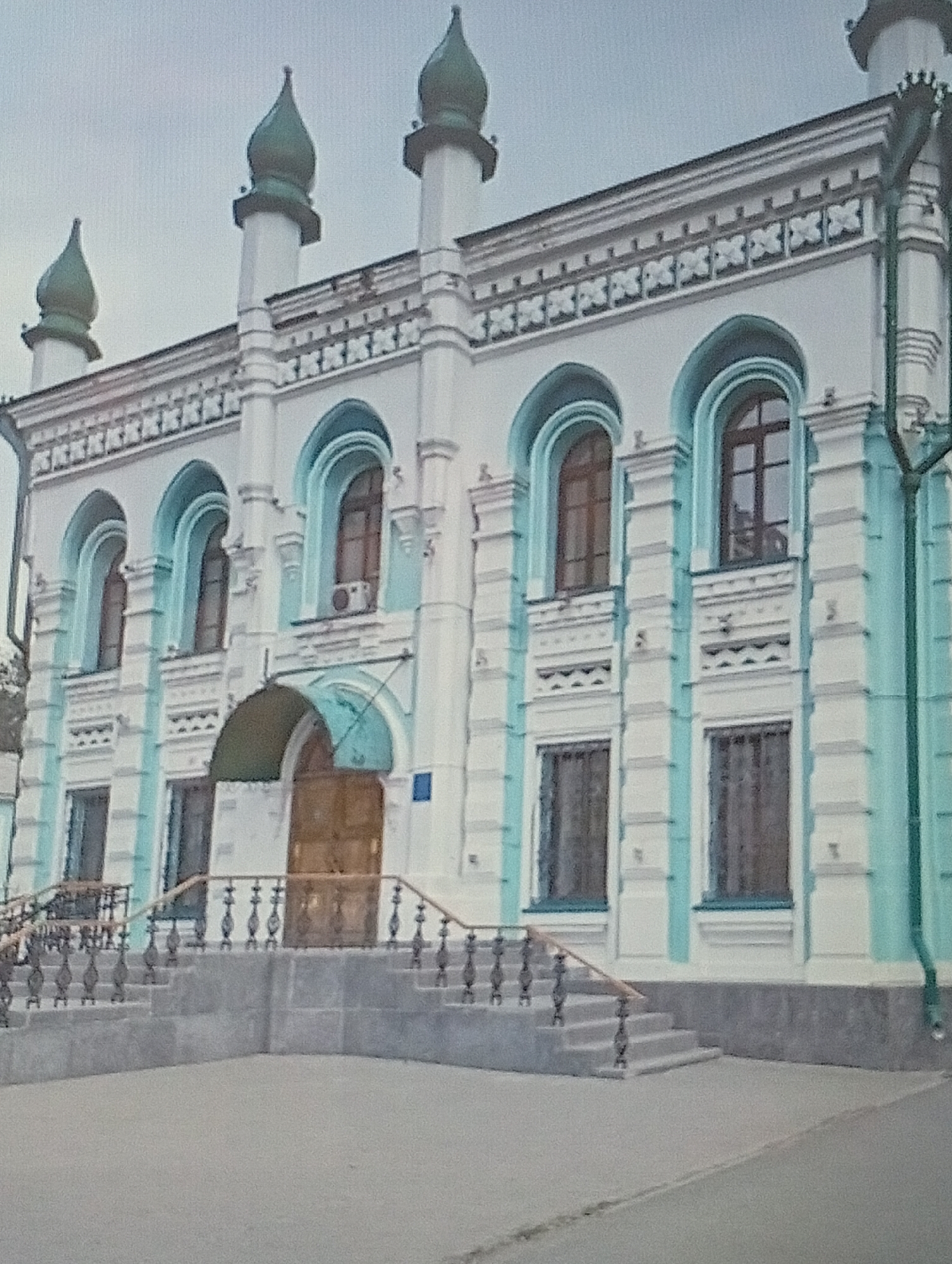 The image shows the exterior of a building in Oral, Kazakhstan, which appears to be a historical or religious structure, possibly a mosque or a synagogue, given its architectural features. The building has a light blue and white facade with decorative elements and arched windows. Three prominent green domes rise from white towers on the roof. The entrance features a wooden door and is accessed via a set of stone stairs with ornate railings. The surrounding area is paved, and the sky is overcast. There are no people visible in the image.