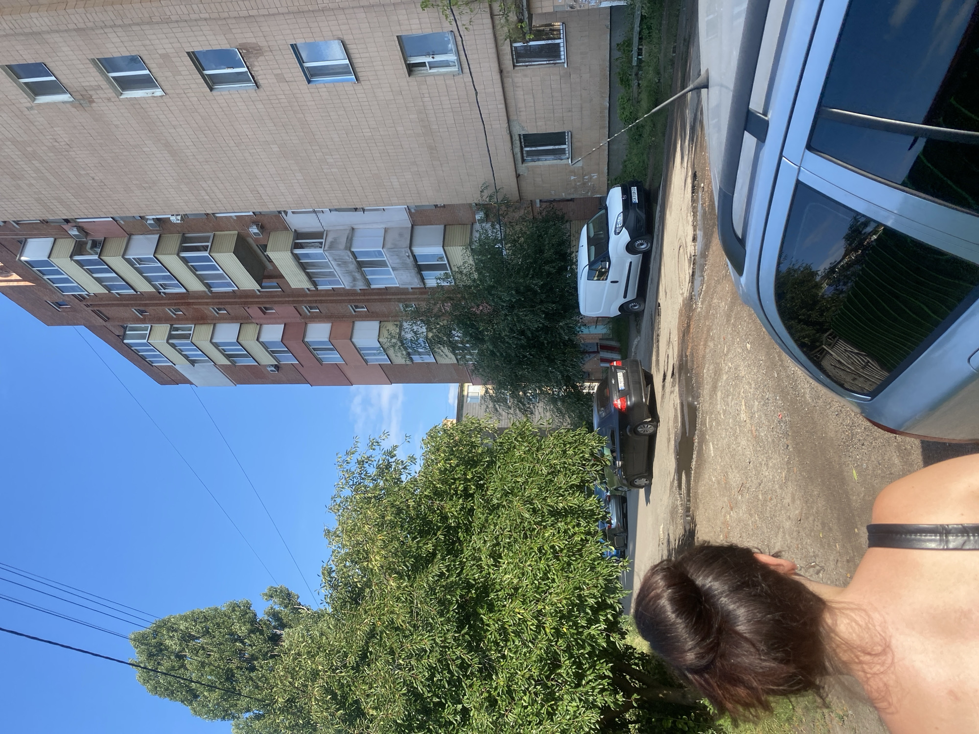 A rear view of a woman with dark brown hair in a bun is captured from the shoulders up, facing away from the camera. She is wearing a black top with a visible strap. The scene is outdoors, likely in a city, with a large, leafy green tree on the left side of the frame.
In the background, there is a multi-story apartment building with a light beige tiled facade on the right, and another apartment building with a reddish-brown brick facade and colorful balconies on the left. Several cars are parked on a dirt and gravel road in front of the buildings, including a black sedan, a silver station wagon, and a white van. The sky is clear and blue. The ground appears to be a mix of dirt, gravel, and some puddles, suggesting recent rain or a damp area. Power lines are visible running across the sky. The environment appears to be a residential area in Poltava, Ukraine.
