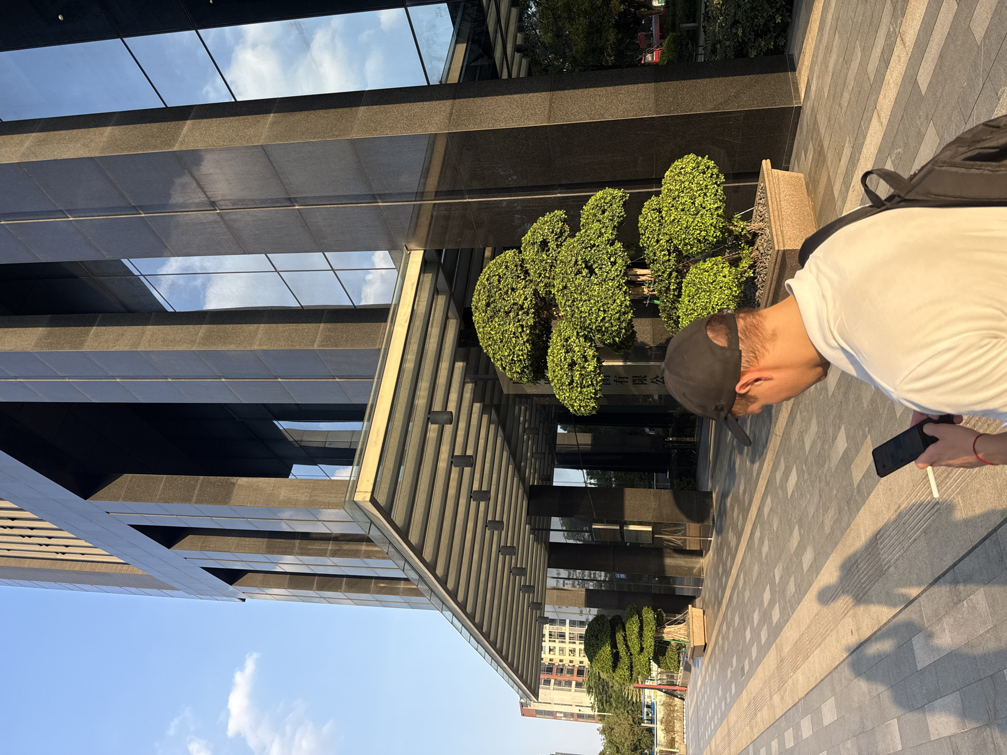 A man with a black baseball cap and white t-shirt is looking at his smartphone, which he holds in his right hand. He has a black backpack on his back. A thin white object, possibly a cigarette or pen, is in his left hand. He stands on a paved walkway. Behind him is a modern glass and dark stone office building. There are pruned bushes planted along the walkway. The building is located in Haizhu District, China, as indicated by the presence of Chinese characters on the building. Other buildings and trees are visible in the distance. The sky is blue with some clouds visible in the glass of the building. The scene is brightly lit by sunlight.