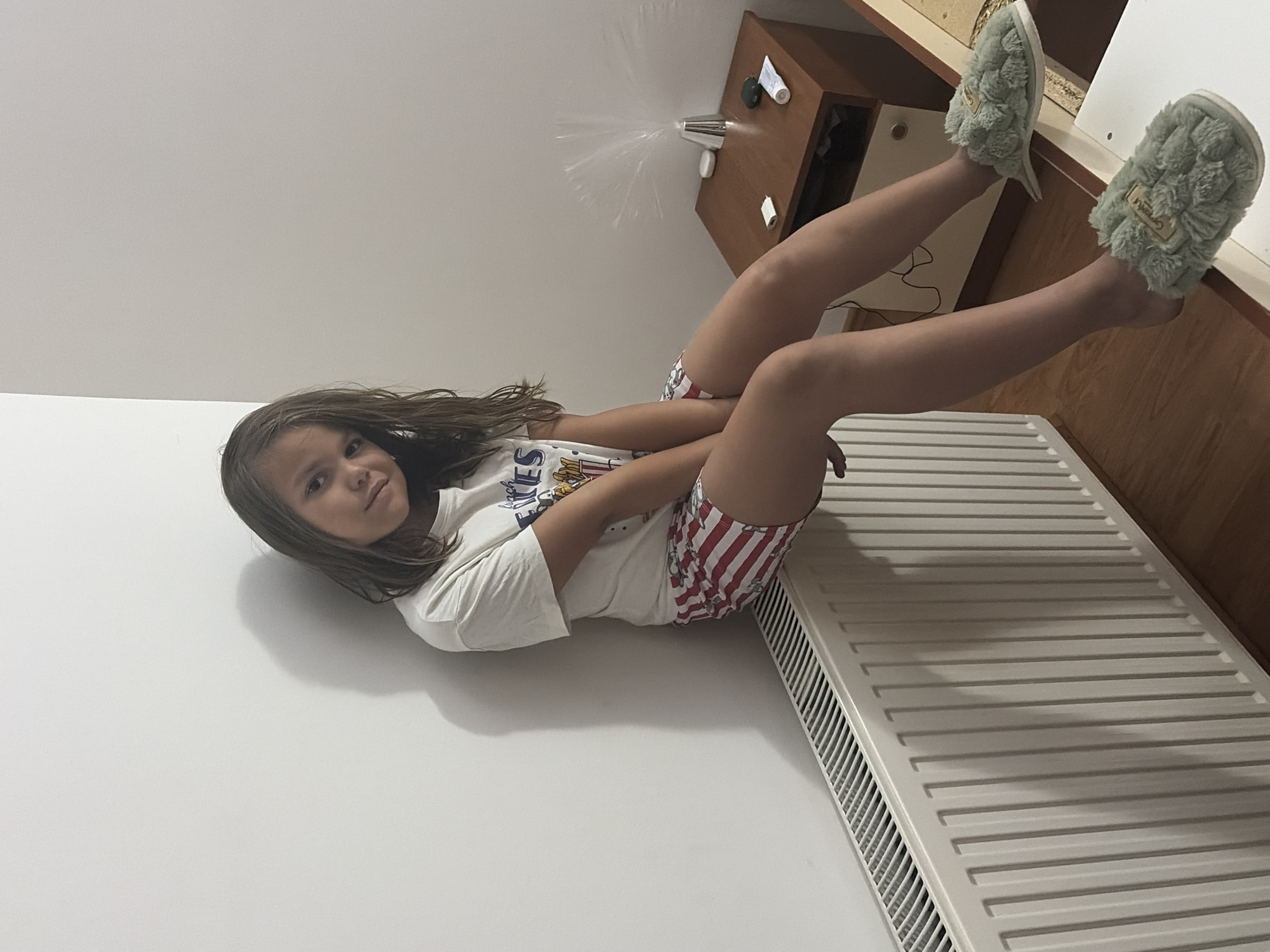 A young girl with long brown hair is lying on a white wall with her legs extended, wearing a white t-shirt and striped shorts. She is also wearing fluffy light green slippers. Her body is positioned as if she is in mid-air, with her legs bent at the knees and her feet pointed upwards. To the right of the girl, a radiator is visible, and behind her, a wooden cabinet with a lamp and some items on top is present. The setting appears to be indoors, possibly a bedroom or living room, with a wooden floor and white walls. The lighting suggests it might be daytime, though the exact time of day is not ascertainable. The girl's expression is relaxed, and the overall mood of the image is playful and carefree. The visible text on her t-shirt is partially obscured and unreadable. The location is Budva, Montenegro.