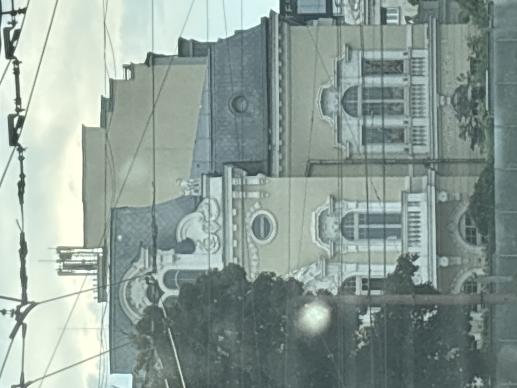 The image shows the facade of an ornate, multi-story building in Sofia, Bulgaria. The building features a mansard roof with intricate detailing, a prominent dormer window, and decorative trim around its windows and balconies. The facade is painted a pale yellow, with white architectural elements. A large tree partially obscures the left side of the building, and overhead wires crisscross the scene, suggesting an urban environment likely with a tram line. The sky is overcast. The building appears to be historical, with a classic architectural style often found in European cities.