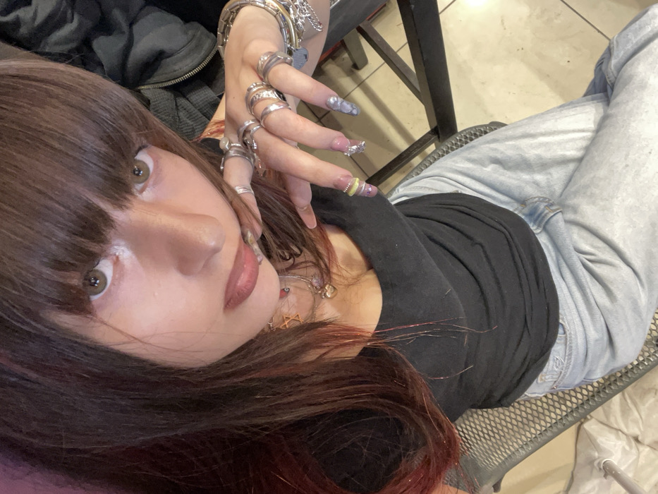 The image presents a close-up, overhead view of a female individual seated indoors. The subject has long, dark brown hair with bangs and reddish tinsel-like highlights, light brown eyes, and is wearing dark lipstick. She is dressed in a black scoop-neck top and light blue denim jeans. Her left hand is raised, prominently displaying numerous silver-toned rings on almost every finger and a layered silver-toned bangle bracelet on her wrist. Her fingernails are long, artificial, and intricately decorated with various designs, including glitter, silver foil, and marbled patterns. Several necklaces are visible around her neck, featuring charms such as a Star of David and a heart-shaped locket. The background is blurred, showing a dark, mesh-patterned chair to the lower left and what appears to be a light-colored tiled floor. The specific location within Raanana, Israel, is not visually discernible.